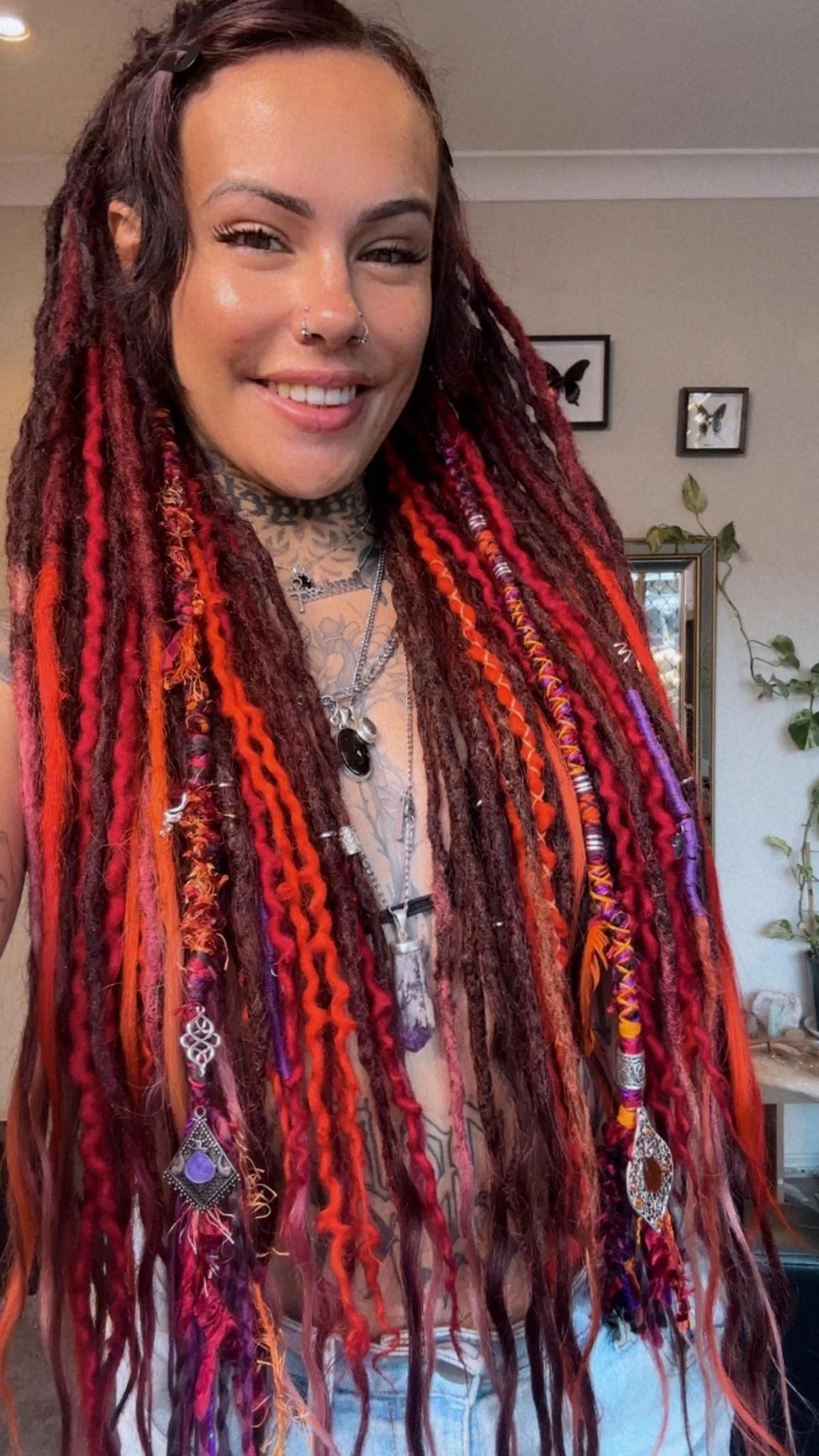A woman with dreadlocks dyed in red, orange, and purple, smiling at the camera, wearing multiple necklaces and earrings, standing in a room with framed butterfly pictures and a mirror in the background.