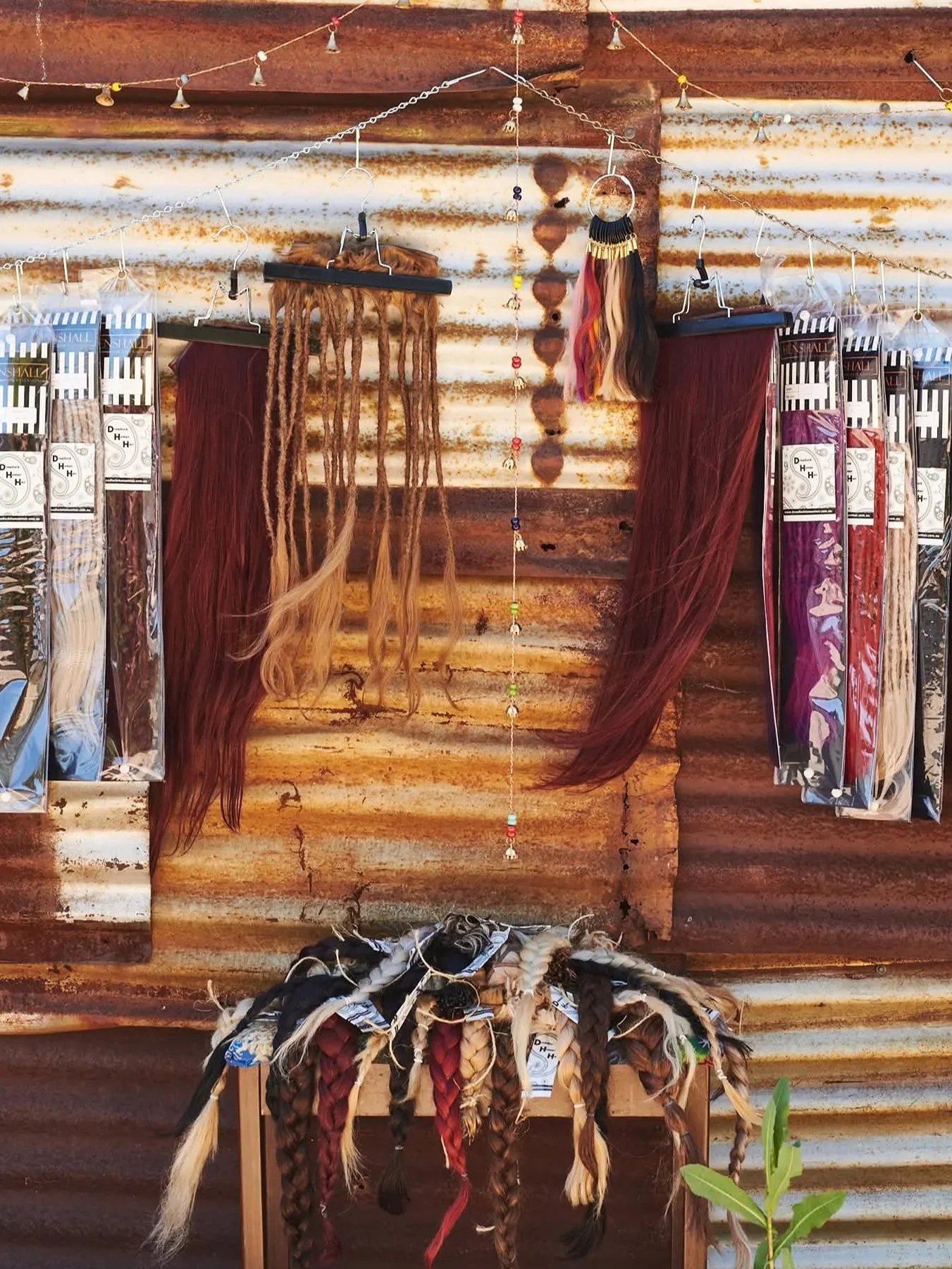 Display of synthetic hair extensions and hairpiece accessories hanging against a rustic, rusty metal wall. The setup includes various braided and straight hairpieces, some with colorful, faux-dreadlock styles, and decorative beads and hooks.