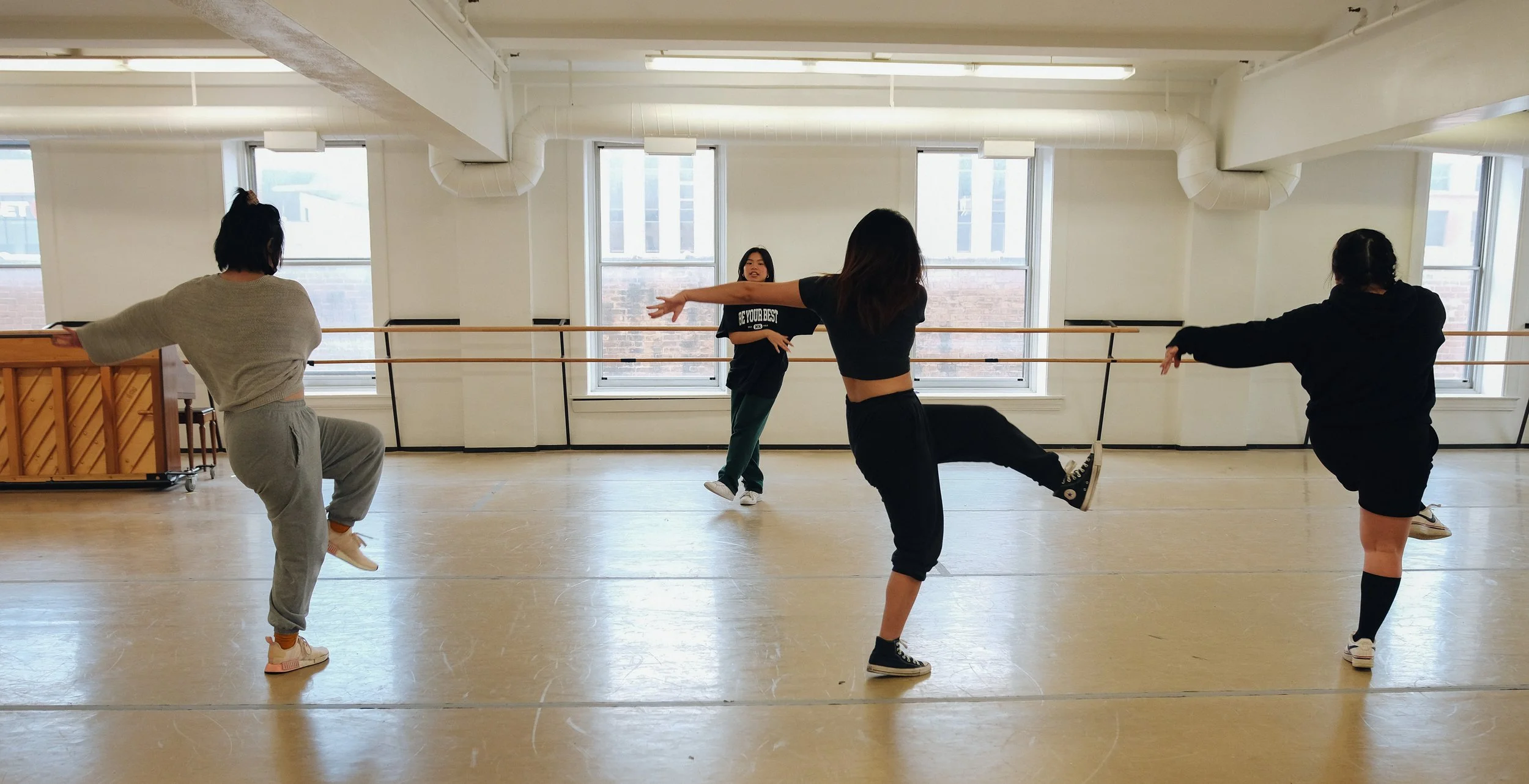 Four women in casual clothing practicing dance moves in a dance studio with large windows and wooden floors.