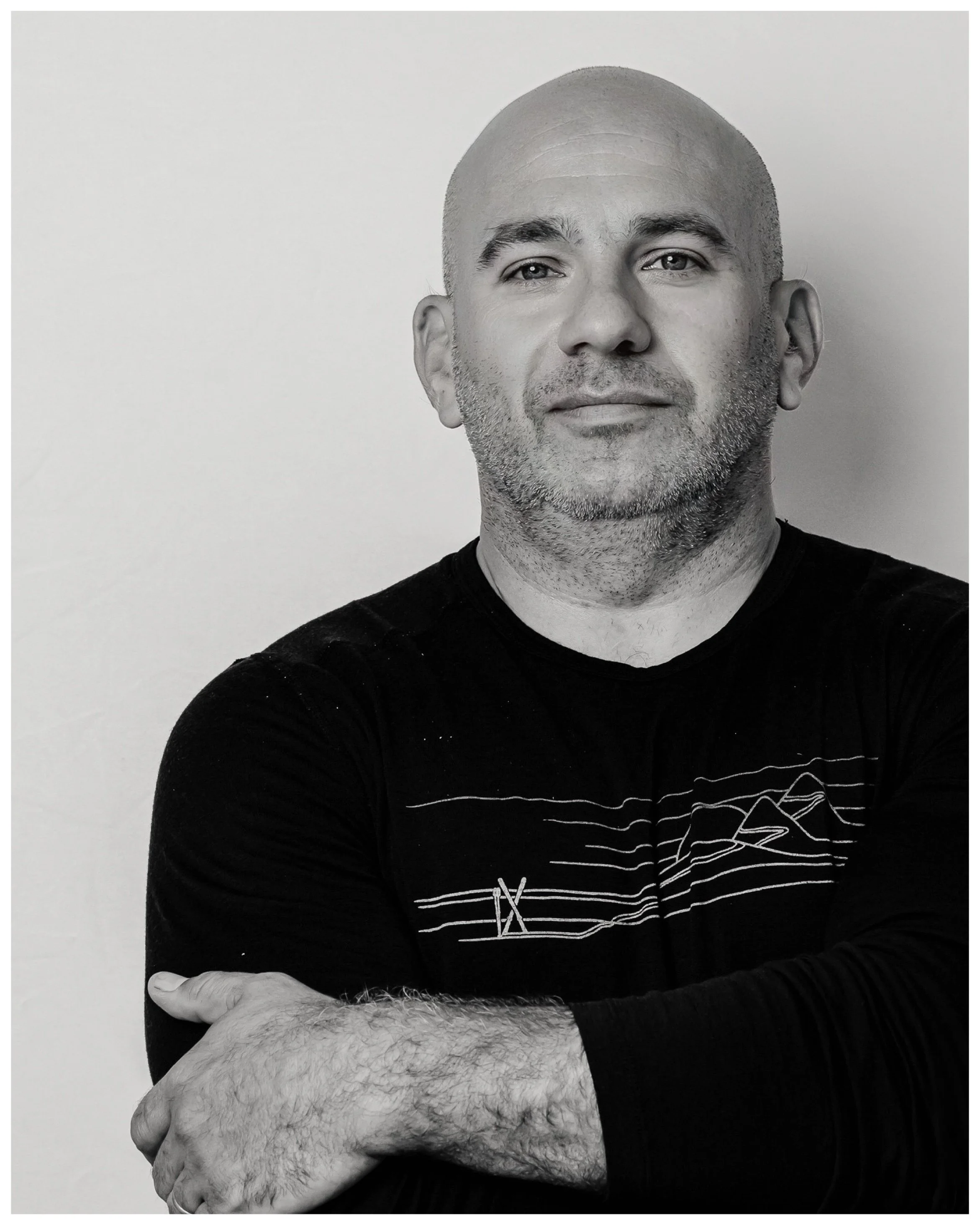 A black-and-white photo of a man with a shaved head and facial hair, wearing a black shirt, standing against a plain light-colored wall. His arms are crossed, showing white hair on his forearms.