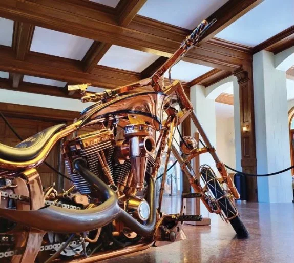 A motorcycle made entirely of shiny, gold-colored metal inside a well-lit room with wooden ceiling beams and white walls.