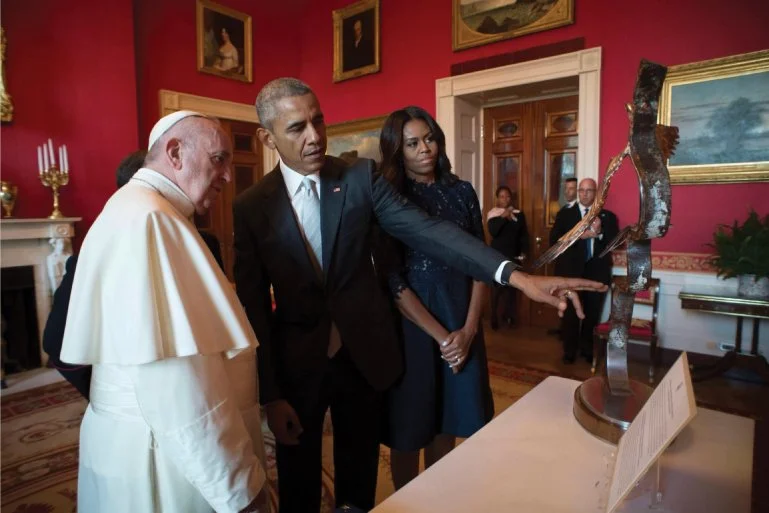 Pope Francis and Barack Obama viewing a sculpture at an art exhibit while Michelle Obama observes, in a richly decorated room with red walls and framed paintings.
