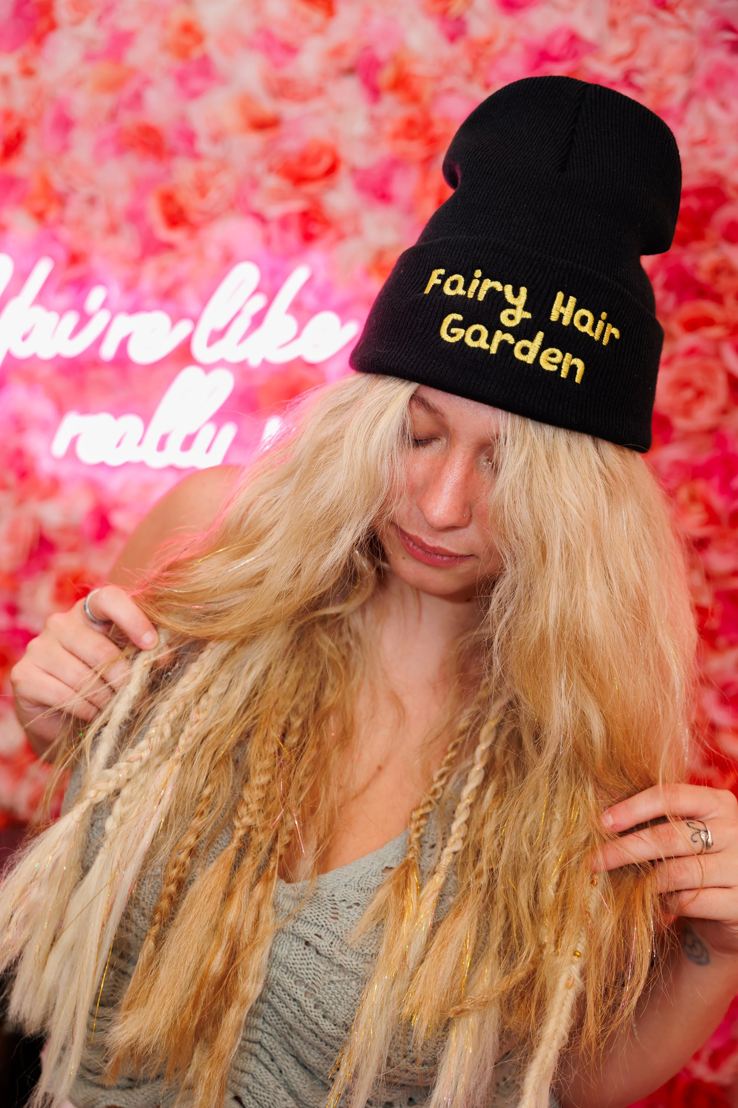 Person with wearing long boho blonde hair wearing a black beanie with 'Fairy Hair Garden' text, surrounded by green leaves and pink flowers.