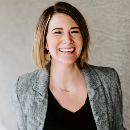 smiling woman with shoulder-length brown hair wearing a gray blazer and black top, against a neutral background