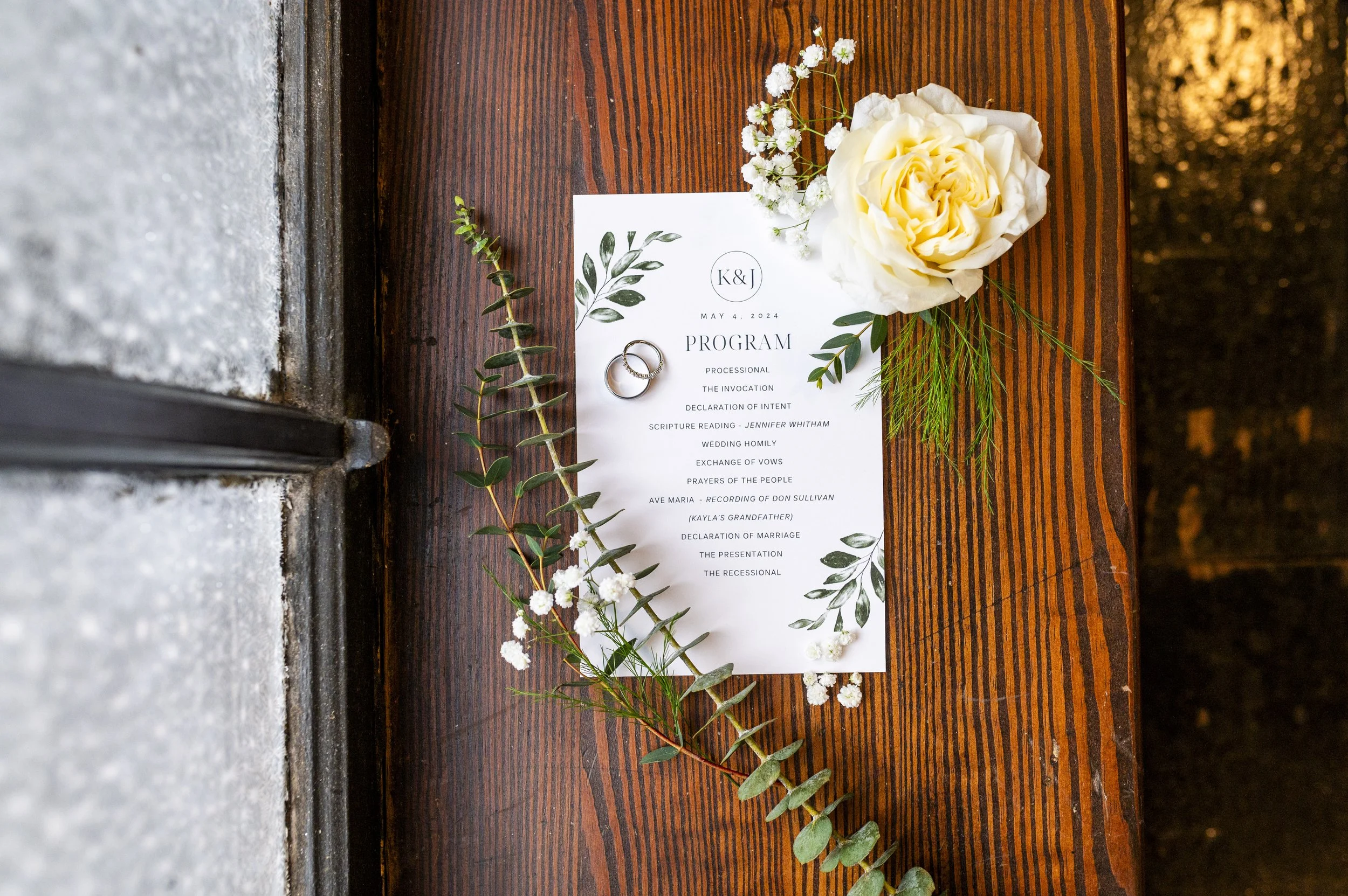 Wedding program on a wooden table decorated with greenery, white flowers, and two wedding rings.