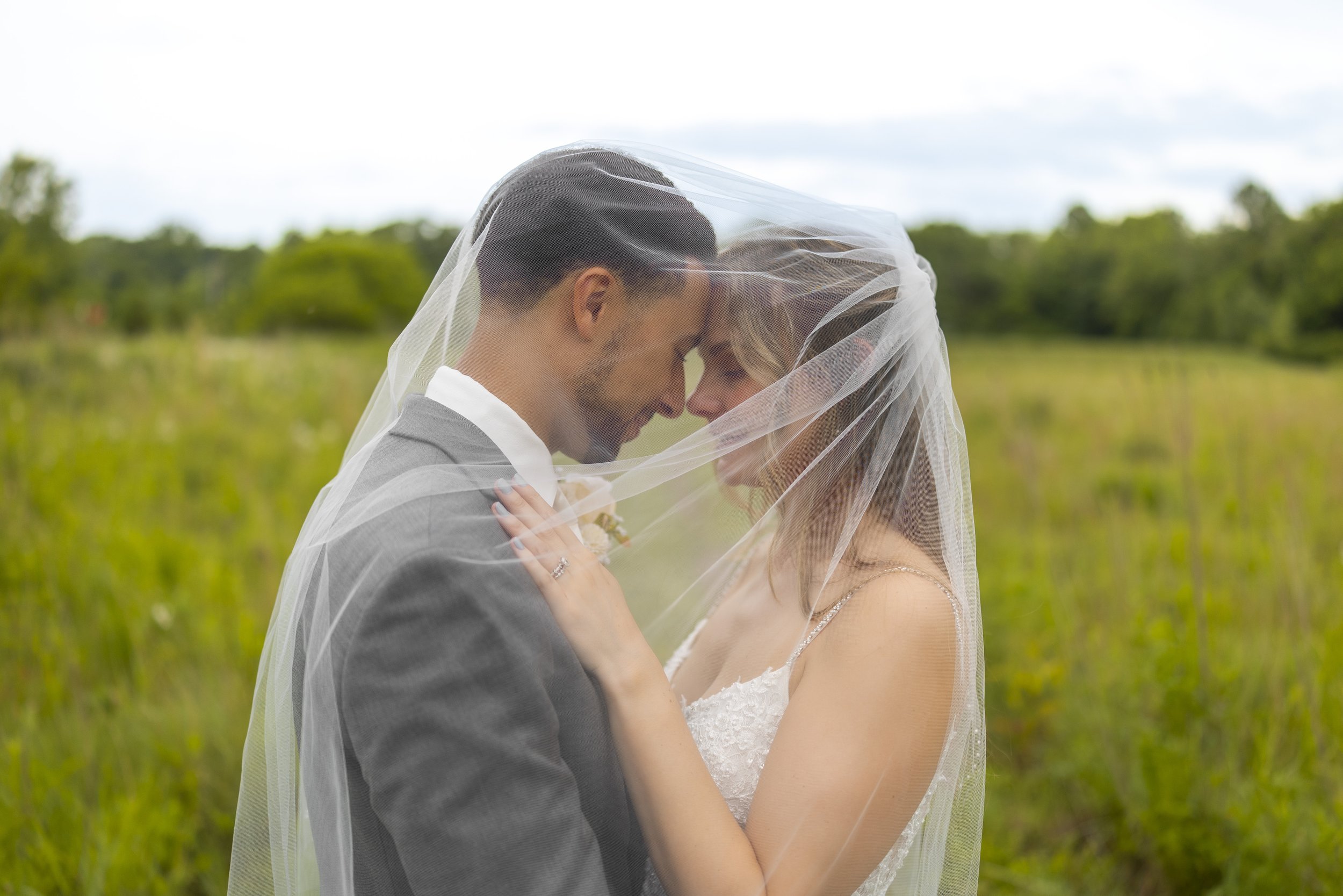 A newlywed couple standing close together outdoors, with their foreheads touching, embracing under a bridal veil in a green field.