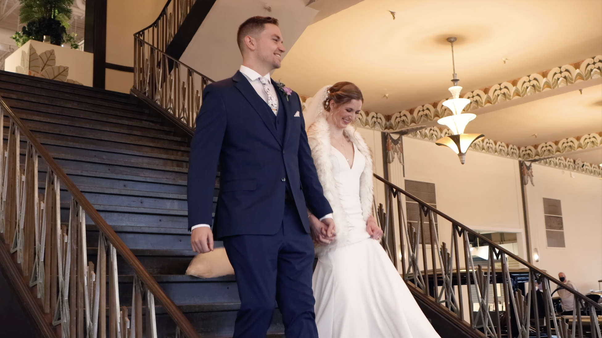 A newlywed couple holding hands and ascending a staircase in a decorated venue, smiling and dressed in wedding attire.