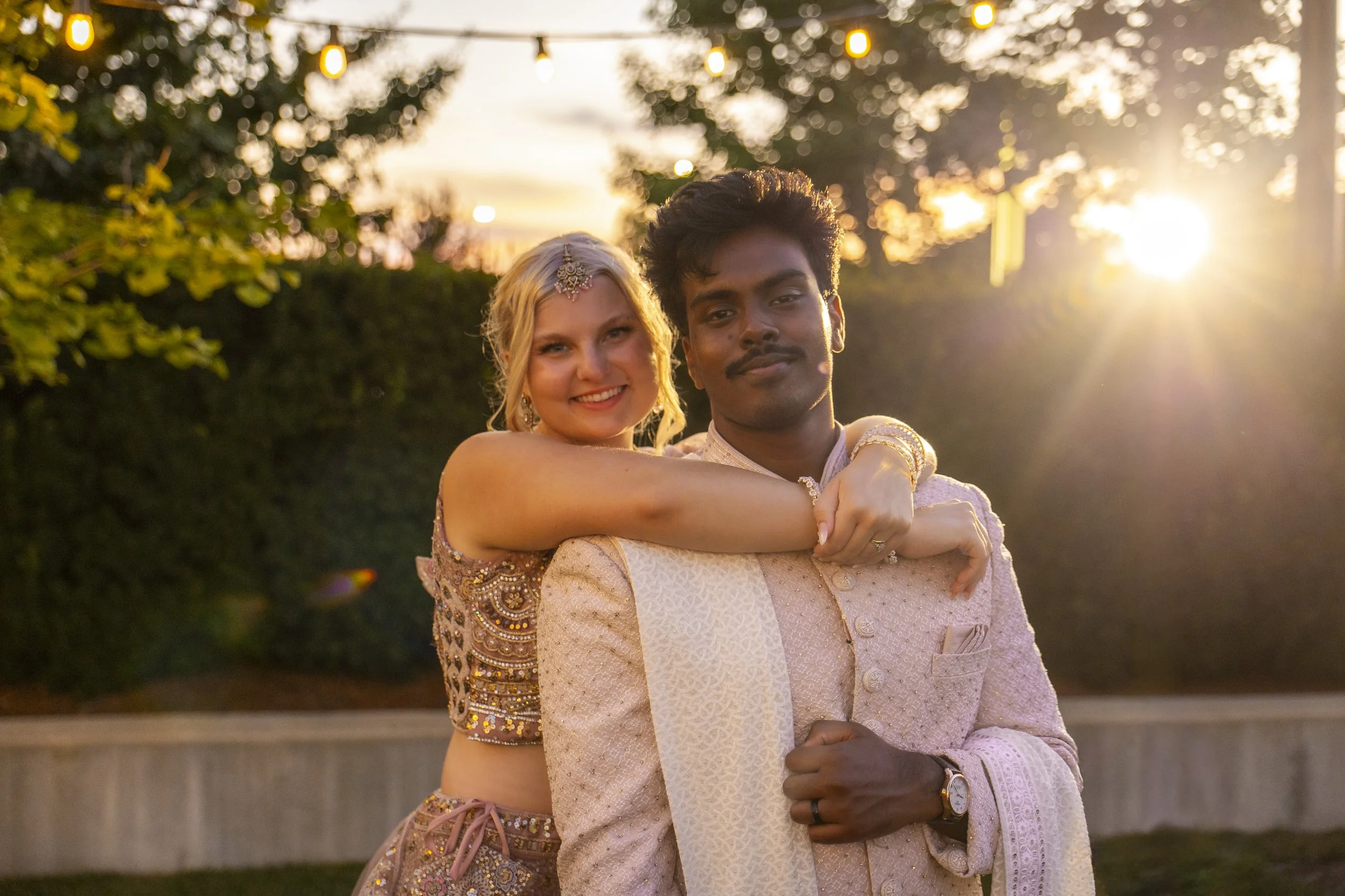 A man and a woman dressed in traditional Indian attire, posing in a garden at sunset with string lights overhead.