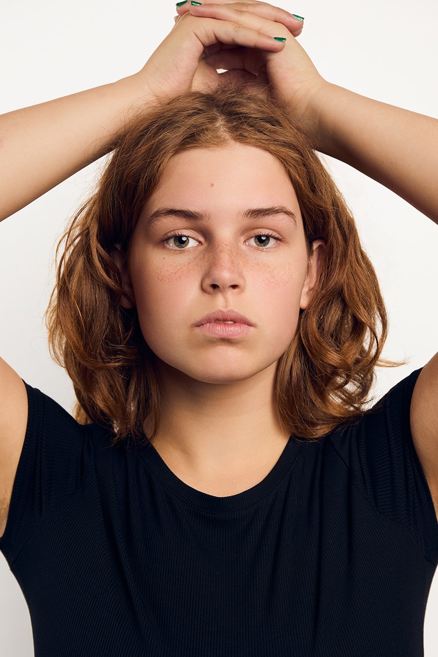 Portrait of a young woman with red hair, freckles, and light skin, wearing a black shirt, with her hands lifted above her head against a plain white background.