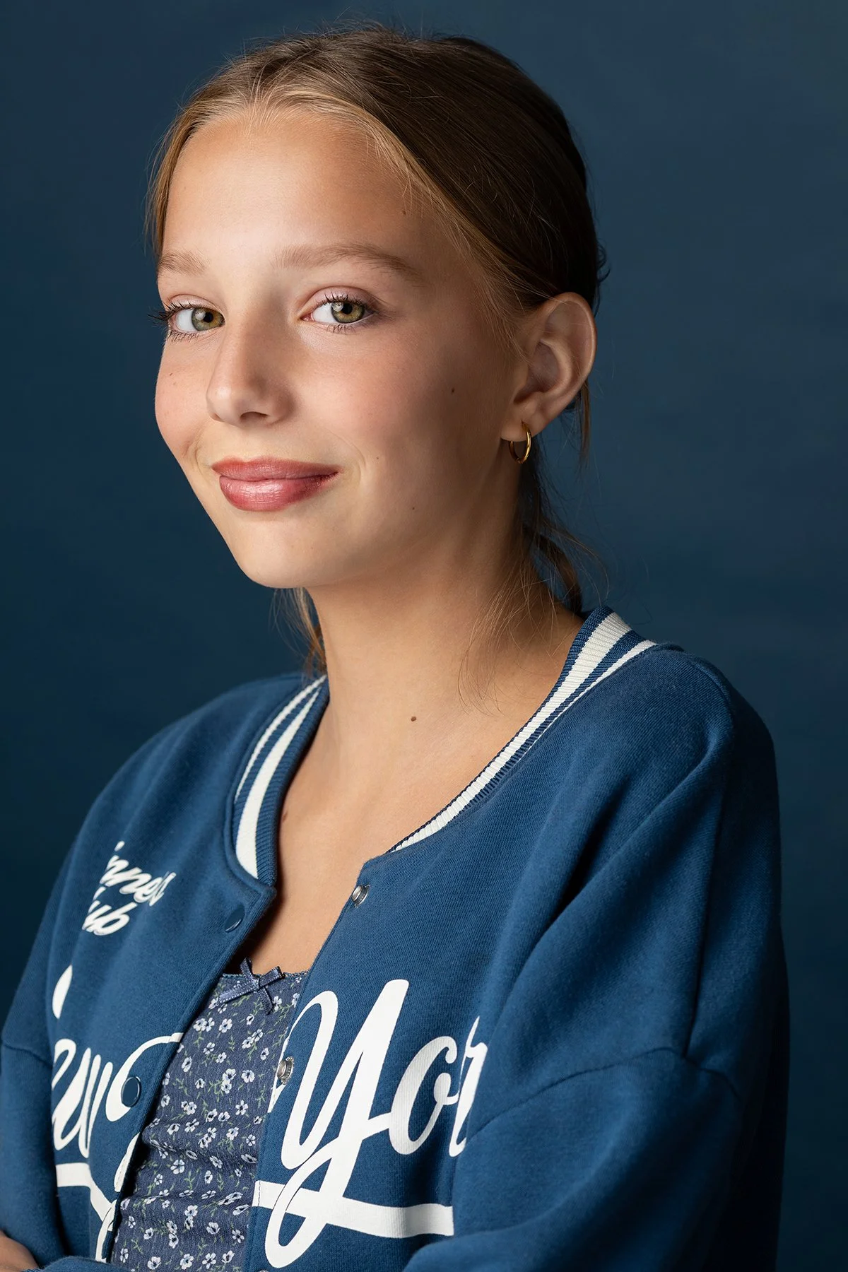 Close-up portrait of a young woman with blonde hair, light skin, and blue eyes, smiling gently, wearing a blue varsity jacket with white lettering and a floral top underneath, against a dark blue background.