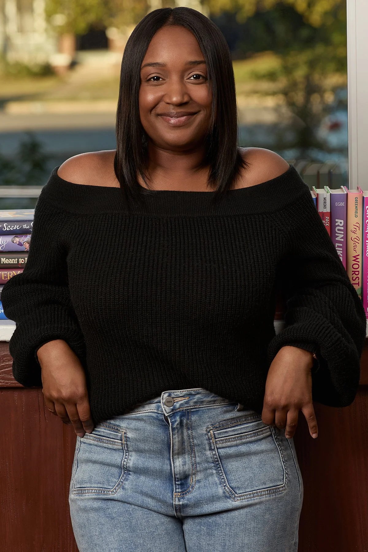 A woman with straight black hair wearing a black off-the-shoulder sweater and light blue jeans, standing in front of a window with a view of trees and buildings outside.