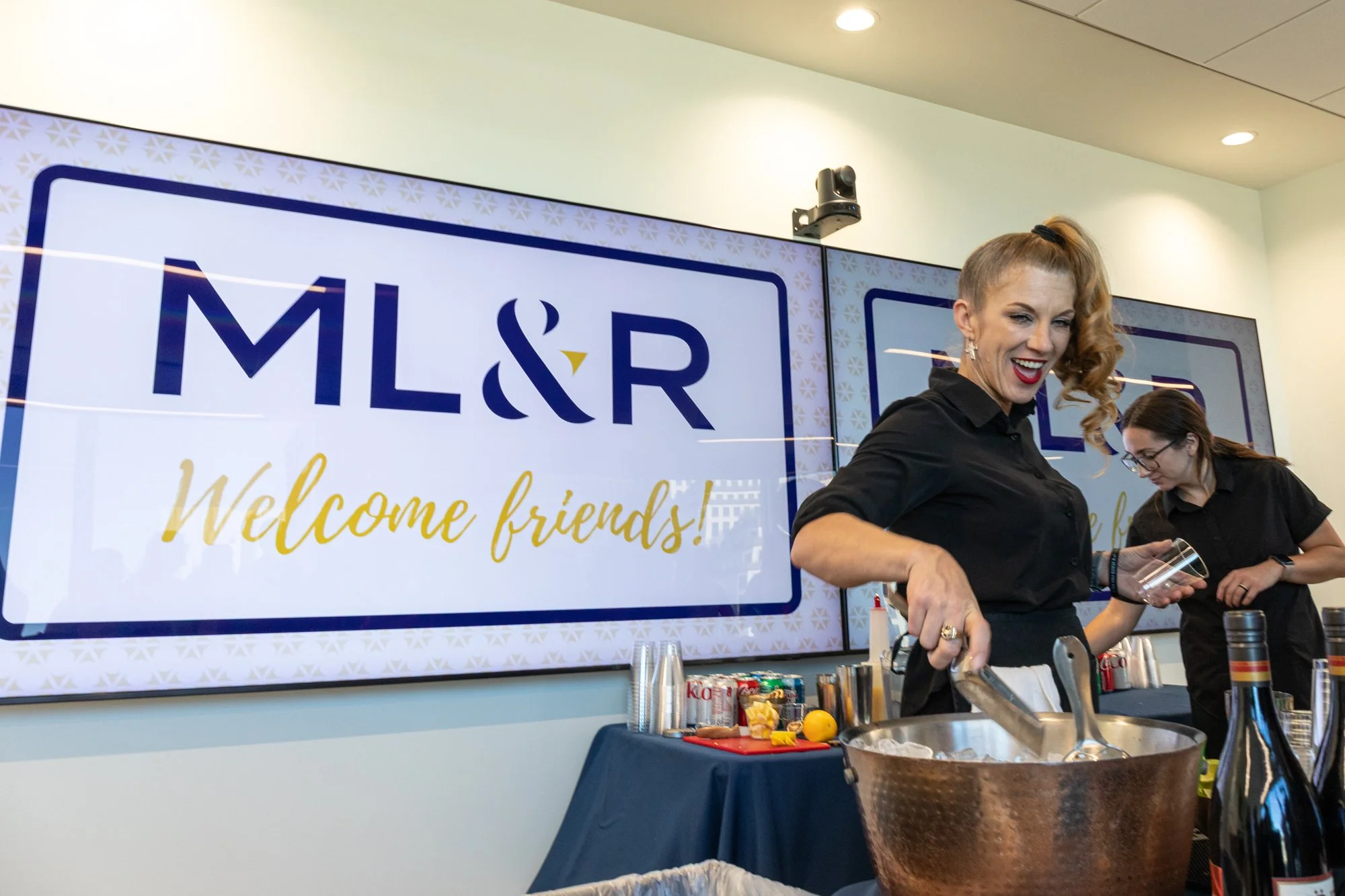 Women at a tasting event with a large screen behind them displaying the text 'ML&R Welcome friends!'.