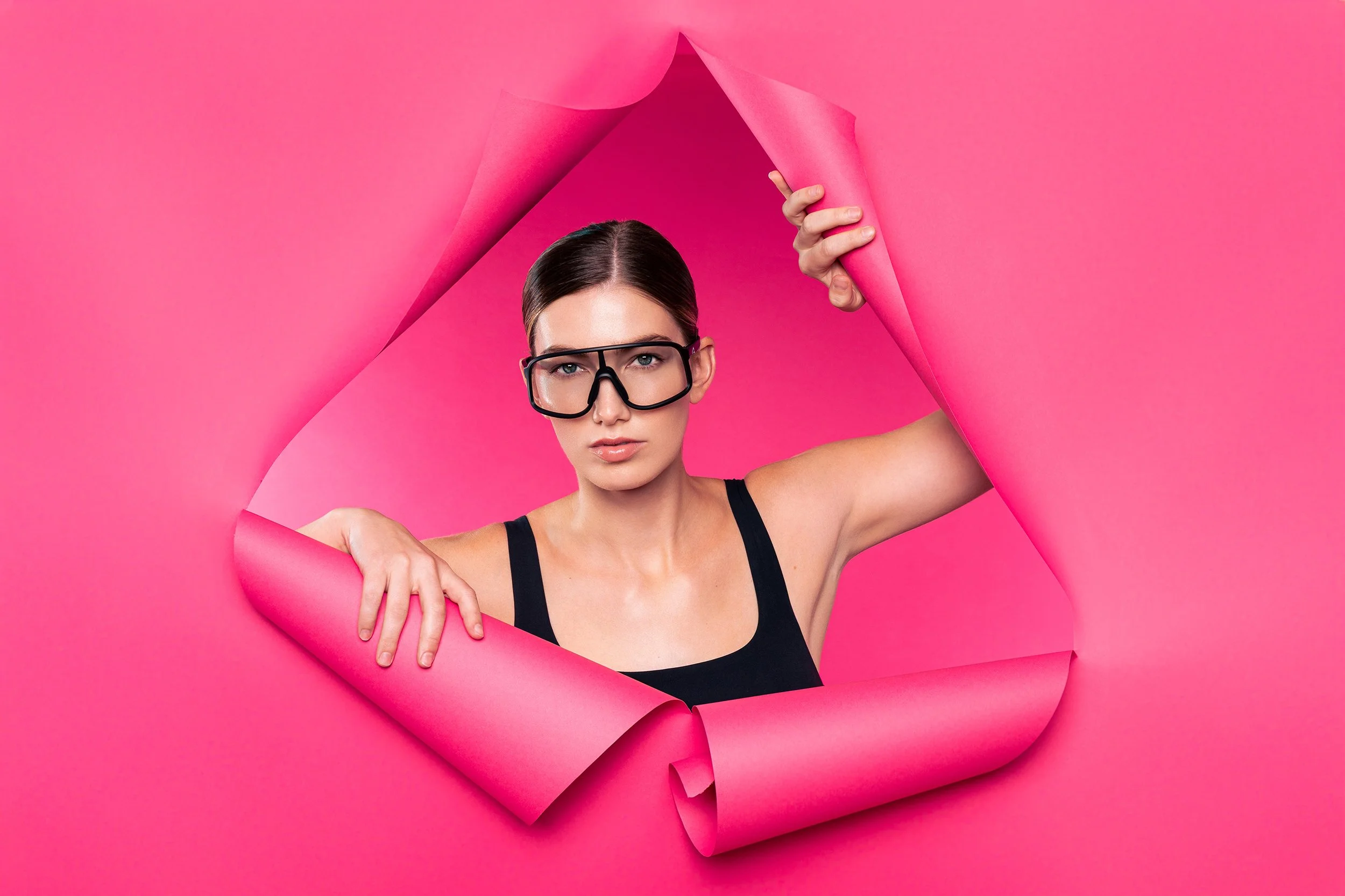 Woman with dark hair, wearing glasses and a black tank top, peering through a pink paper tear in a pink background.