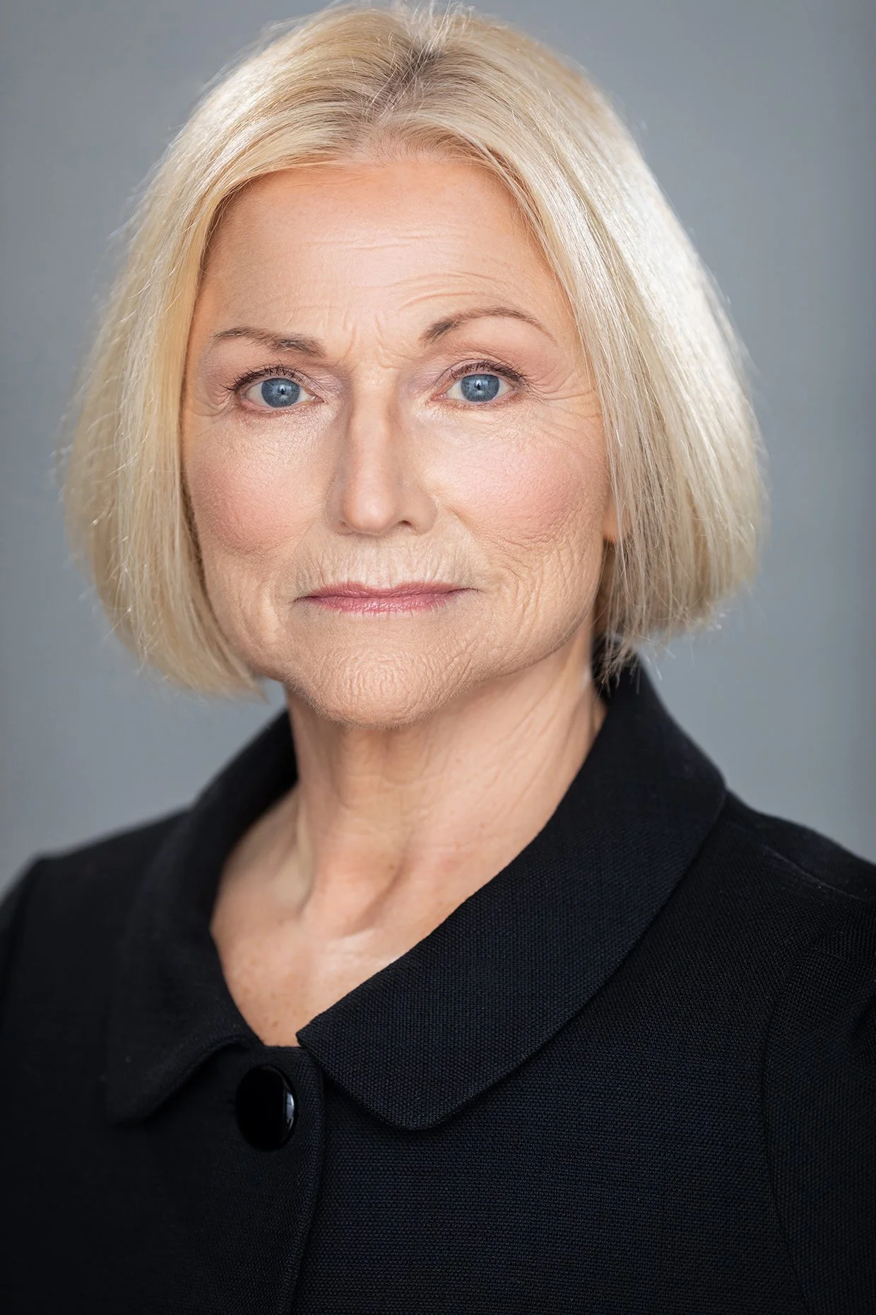 A close-up portrait of an elderly woman with blonde hair, blue eyes, and a serious expression, wearing a black collared top against a neutral gray background.