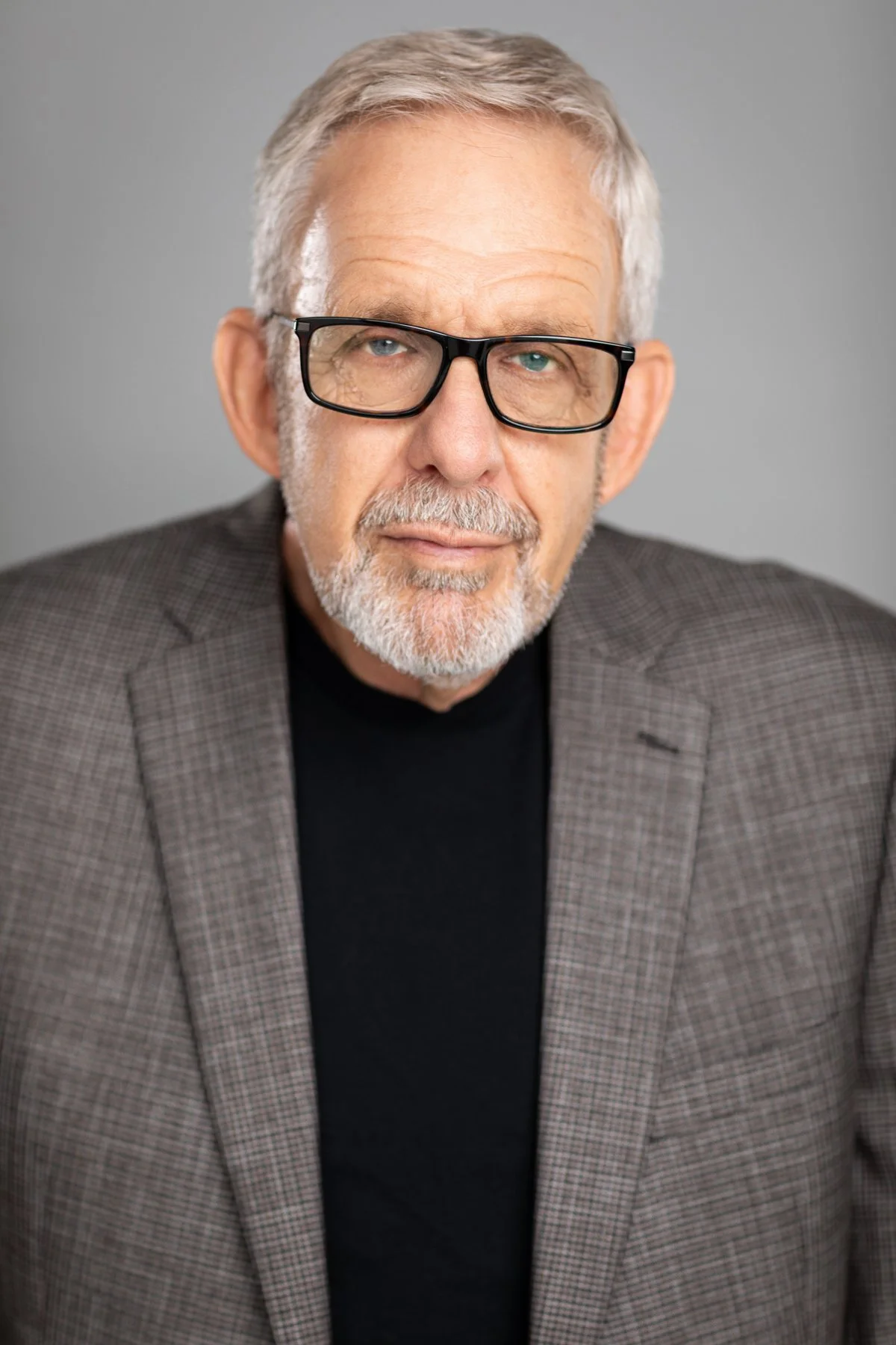 An older man with gray hair, glasses, and a beard wearing a checkered blazer and black shirt, looking thoughtfully at the camera against a plain gray background.