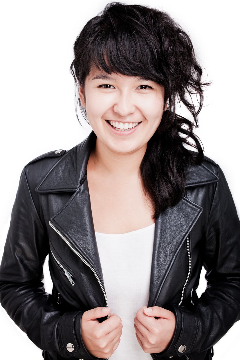 A young woman with dark, wavy hair smiling, wearing a black leather jacket over a white top, against a plain white background.