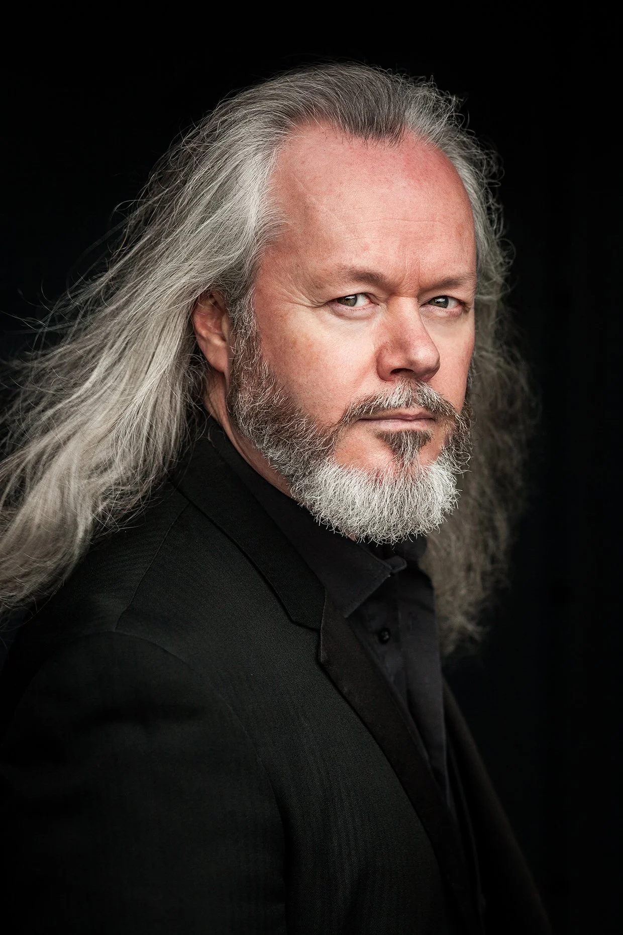 A portrait of a man with long gray hair and a beard, wearing a black jacket, against a black background.