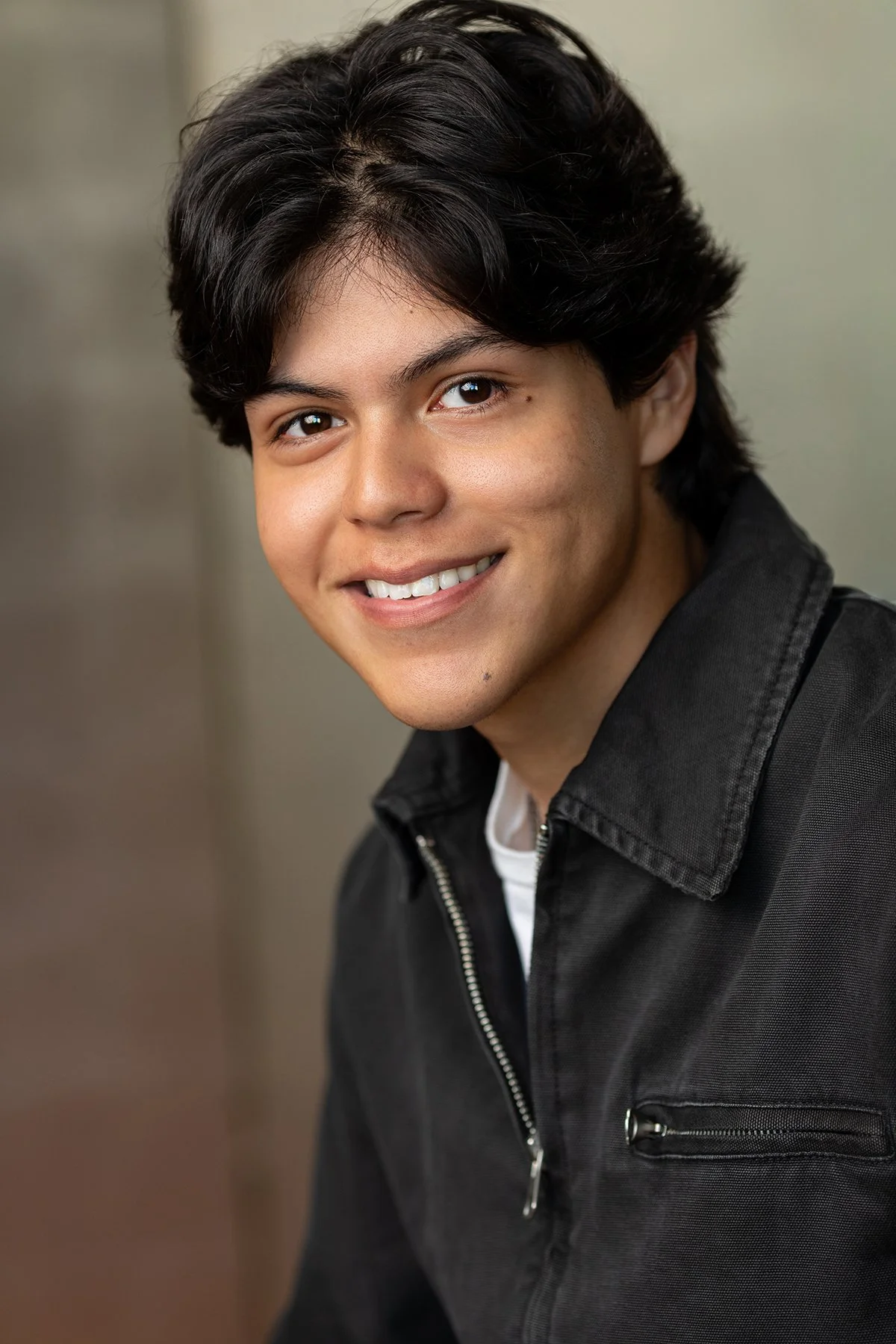 Young man with dark hair smiling, wearing a black jacket, in an indoor setting.