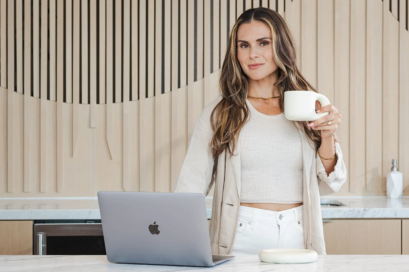 A woman with long brown hair in a beige cardigan and white outfit holding a white mug in a modern kitchen with a grey MacBook on a white countertop.