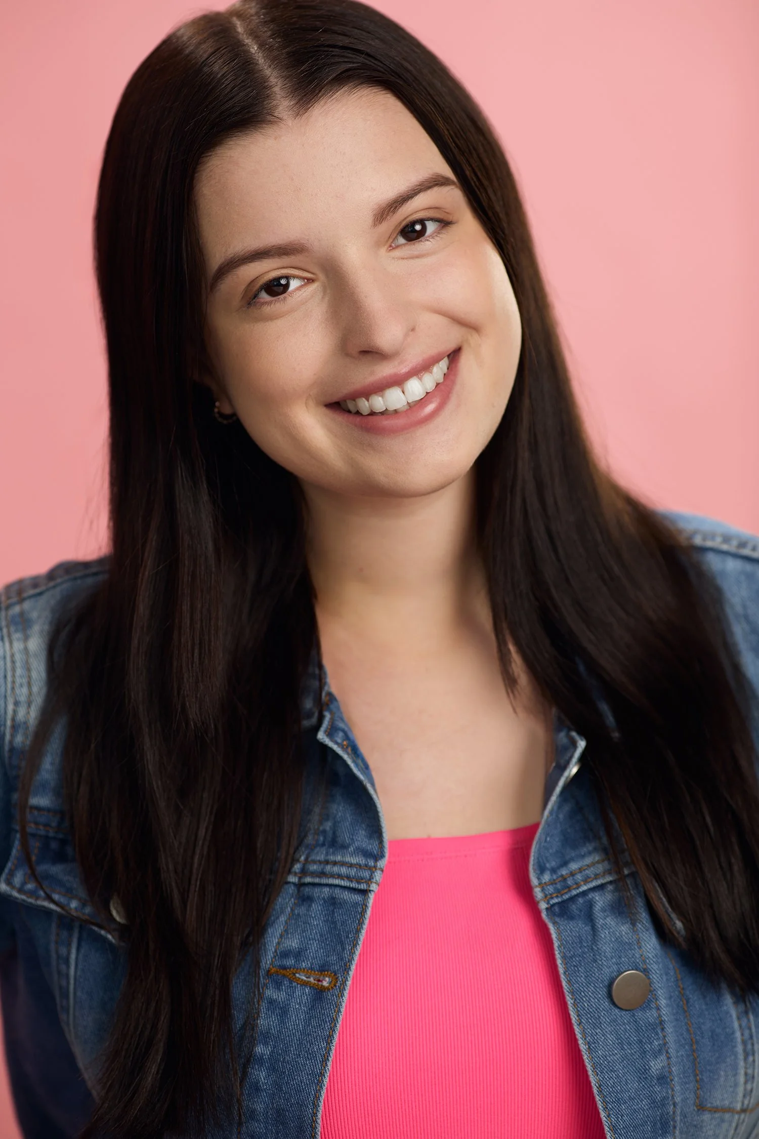 A young woman with long dark hair smiling, wearing a denim jacket over a pink top, against a pink background.