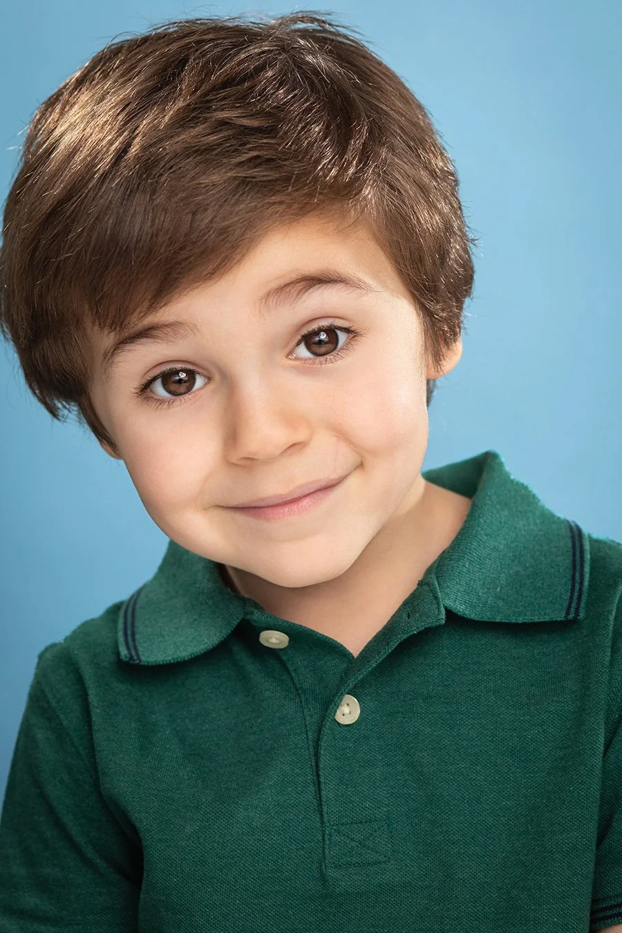 A young boy with brown hair and brown eyes smiling, wearing a green polo shirt, posed against a blue background.