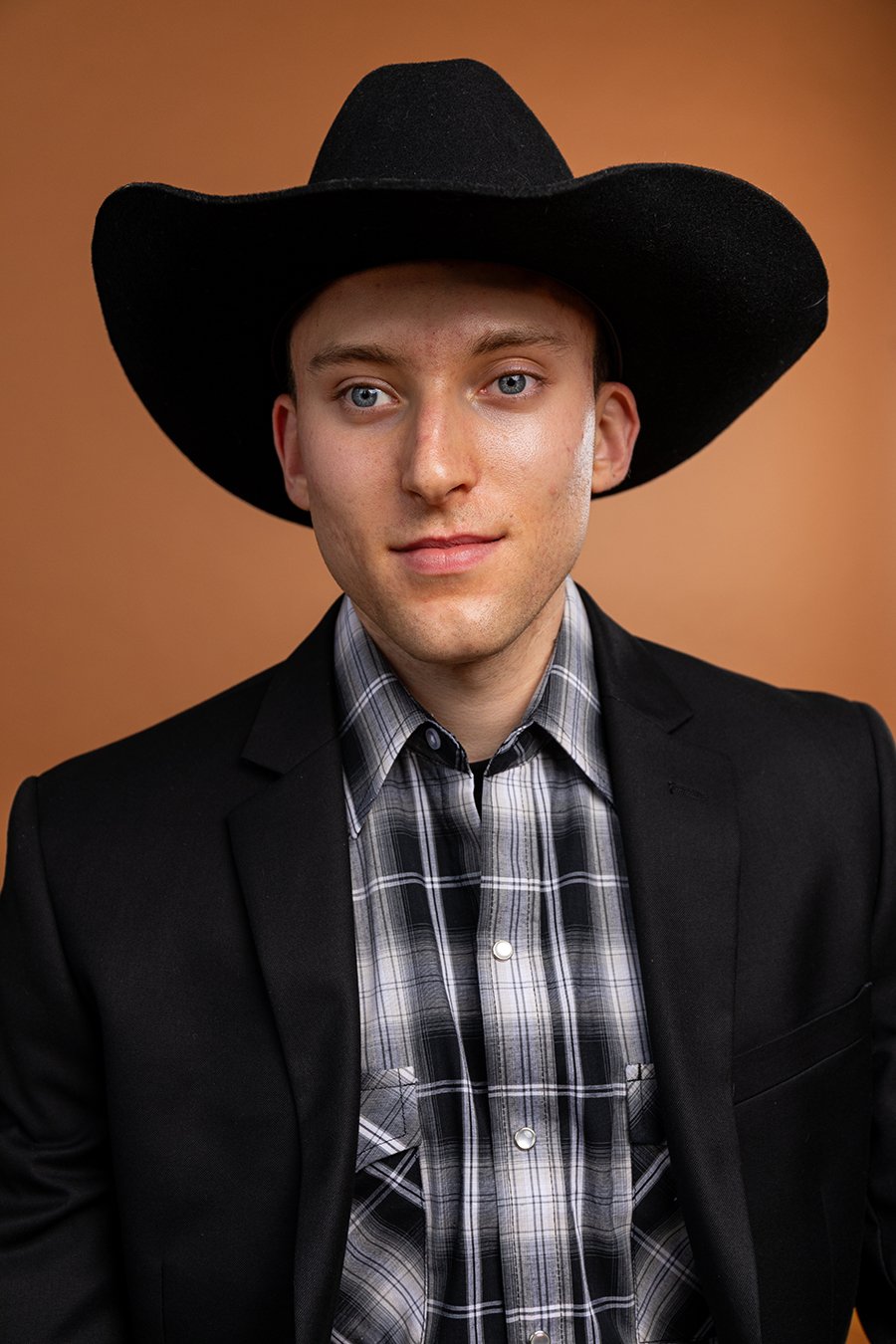 A young man with light skin, blue eyes, and short dark hair wearing a black cowboy hat, a black blazer, and a plaid shirt against a plain beige background.