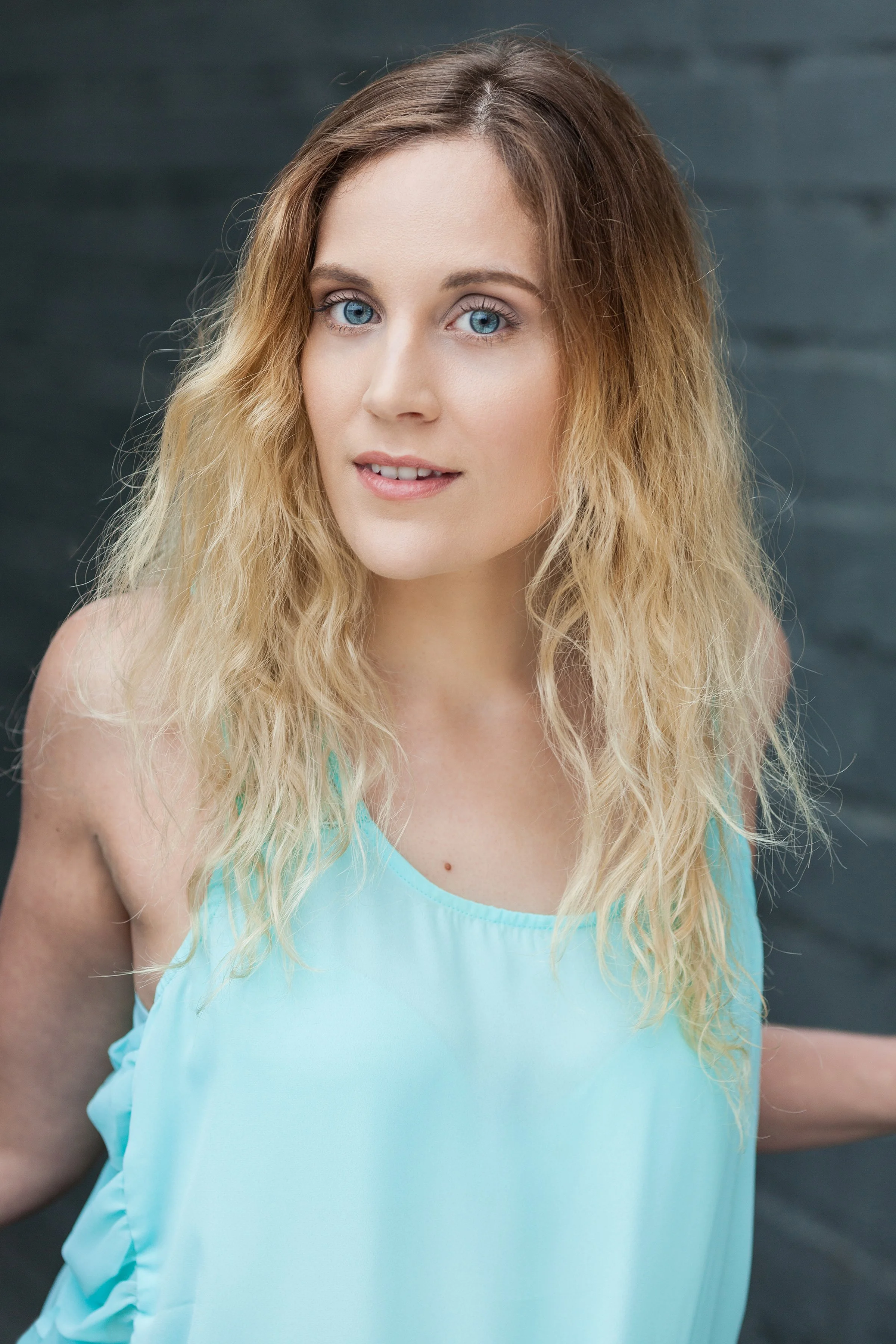 A young woman with long, wavy blonde hair and blue eyes, wearing a light blue sleeveless top, standing in front of a dark textured background.