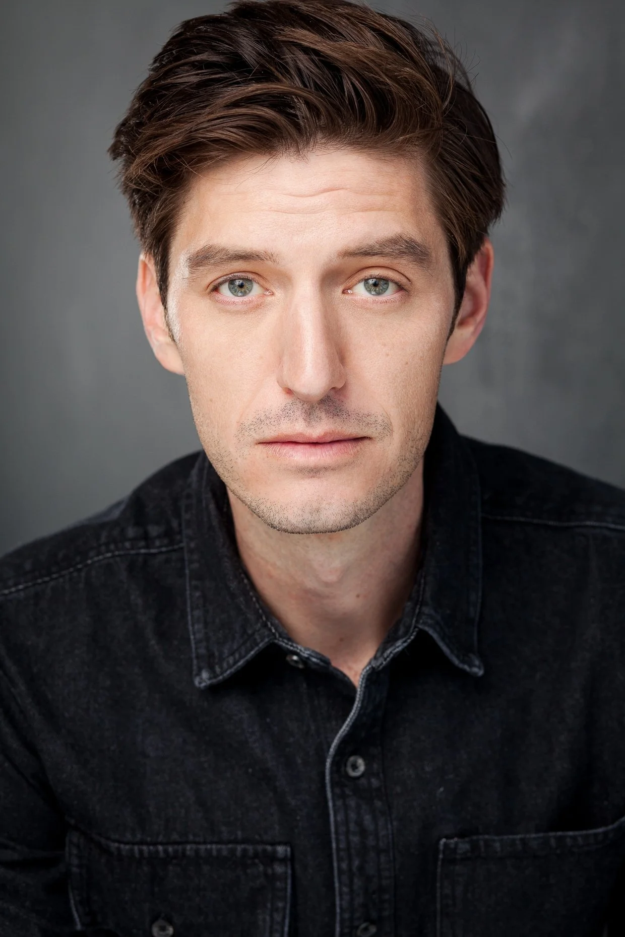 Close-up portrait of a young man with fair skin, brown hair, and blue eyes, wearing a black denim shirt, against a gray background.