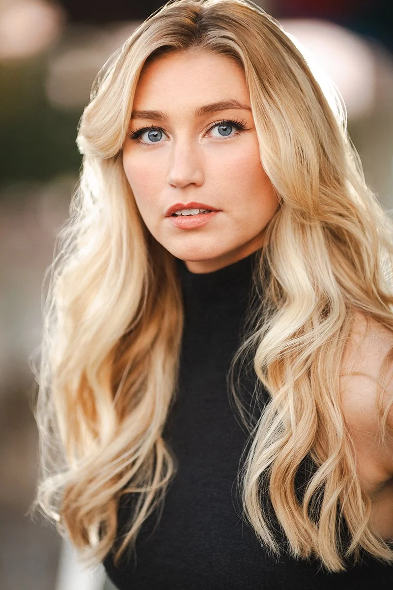 A close-up of a woman with long, wavy blonde hair and blue eyes, wearing a black top, outdoors with blurred background.