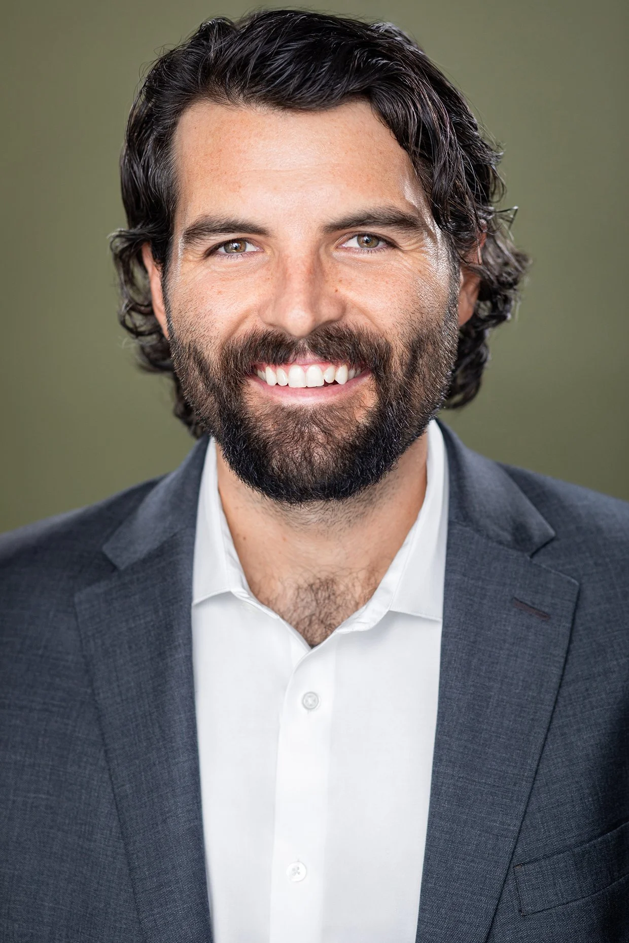 Headshot of a man with dark, wavy hair and a beard, wearing a dark suit jacket and white shirt, smiling against a blurred green background.