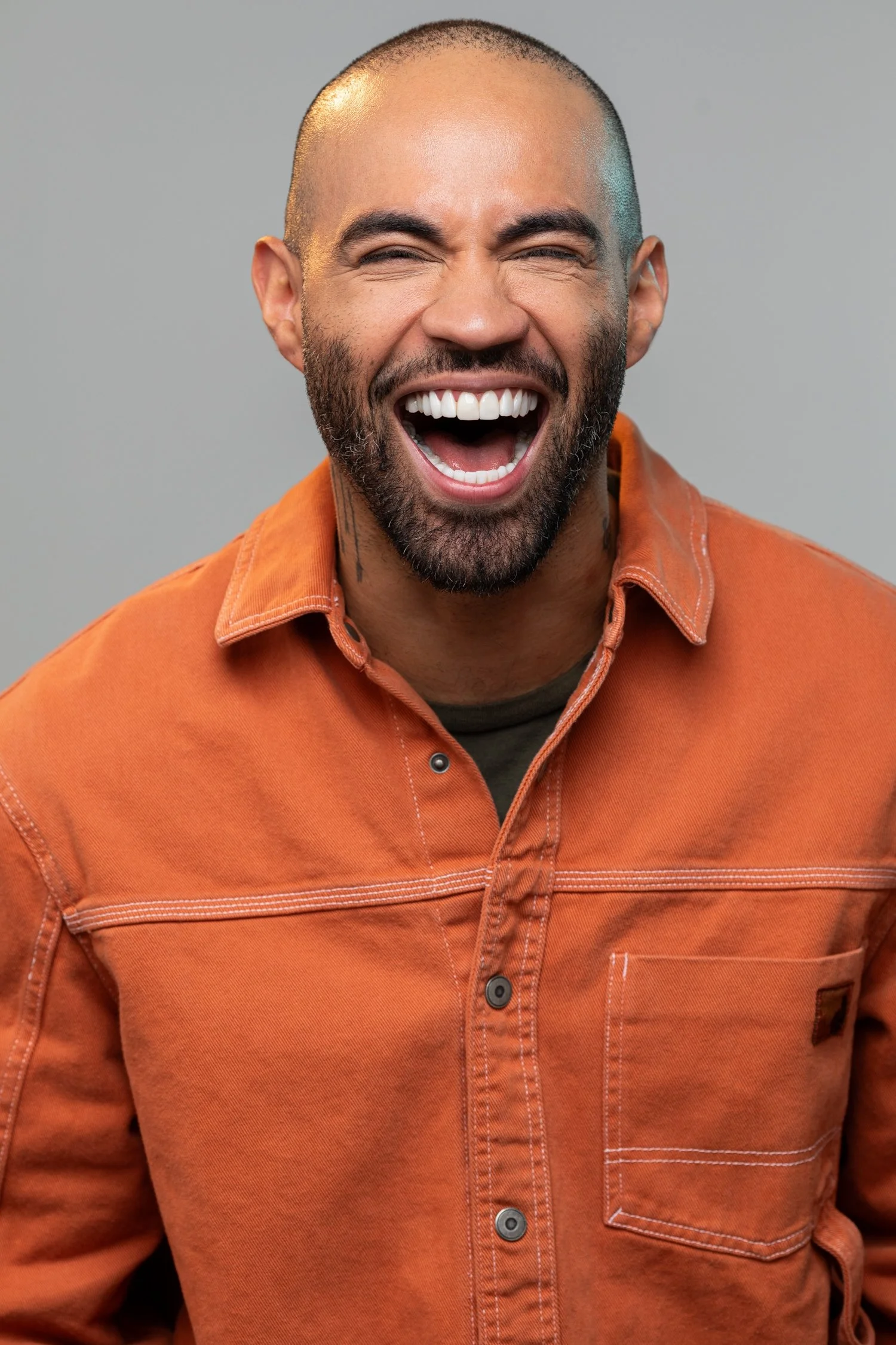 A man laughing happily, wearing an orange jacket, with a gray background.