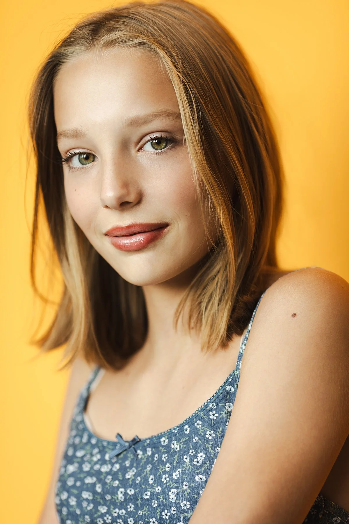 A young woman with wavy, shoulder-length brown hair and light skin, wearing a black V-neck top, standing in front of a wooden wall background.