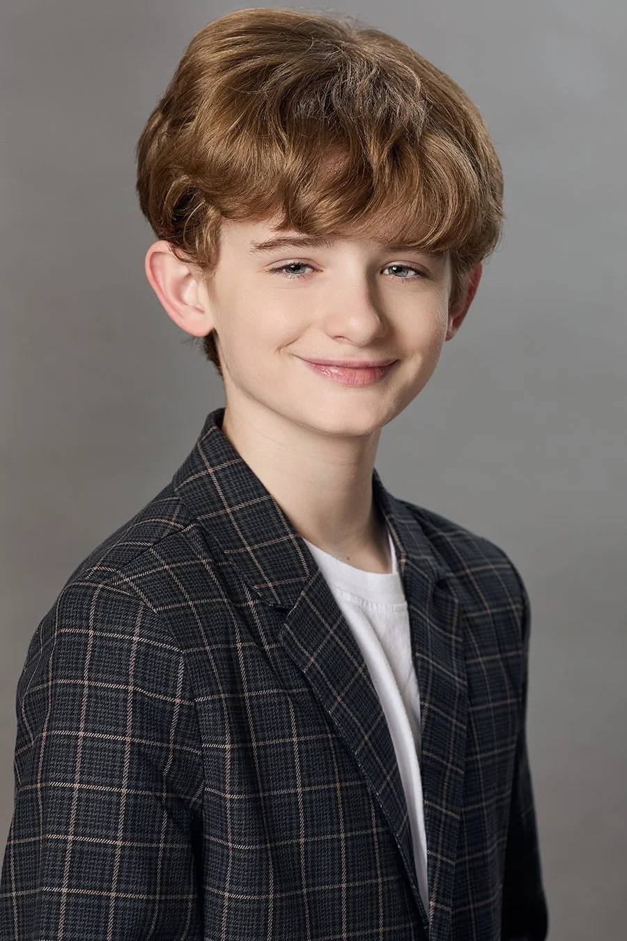 Smiling young boy with light skin, wavy red hair, wearing a black plaid blazer and a white shirt, standing against a gray background.