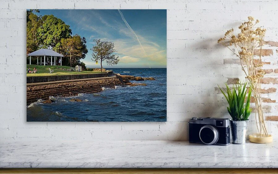A printed photo of a seaside park scene with trees, a gazebo, and a body of water hangs on a white brick wall. On the table below, there is a camera, a vase with flowers, and a decorative plant.