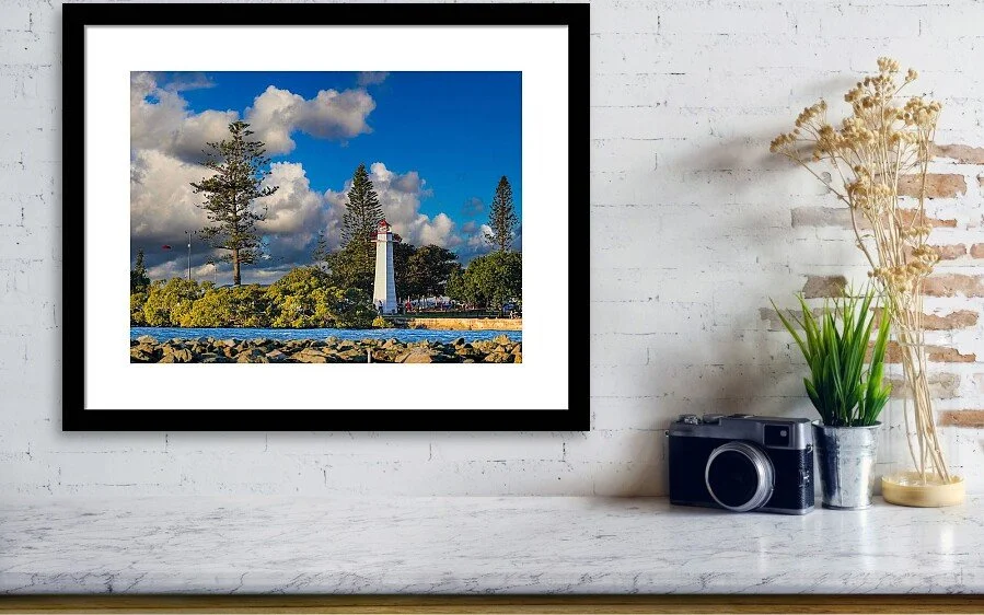 Photograph of a lighthouse and trees on a sunny day with a partly cloudy sky, displayed in a black frame on a white wall