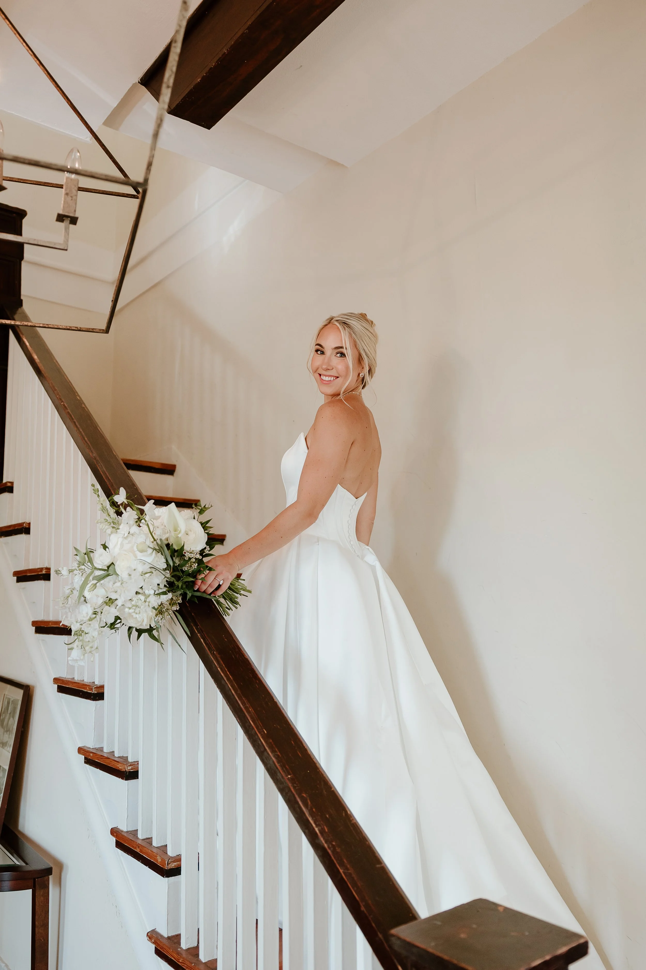 A bride looking at herself in a mirror, wearing a white wedding dress and veil, with pearl jewelry, in a decorated room.