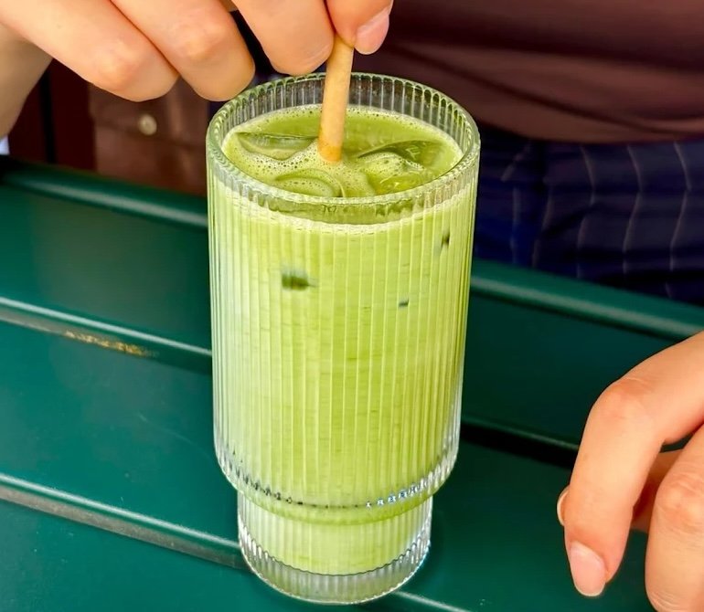 Healthy-looking green drink in glass cup on green table being stirred with straw by female hand.