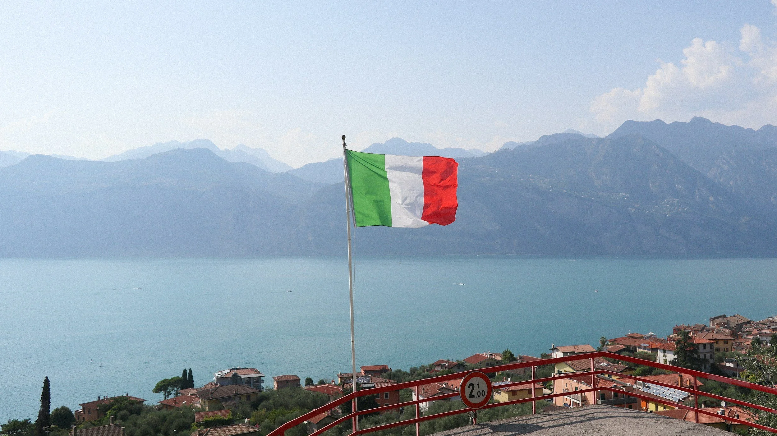 Italian flag waving in wind with mountains, sea, and sky in backdrop.