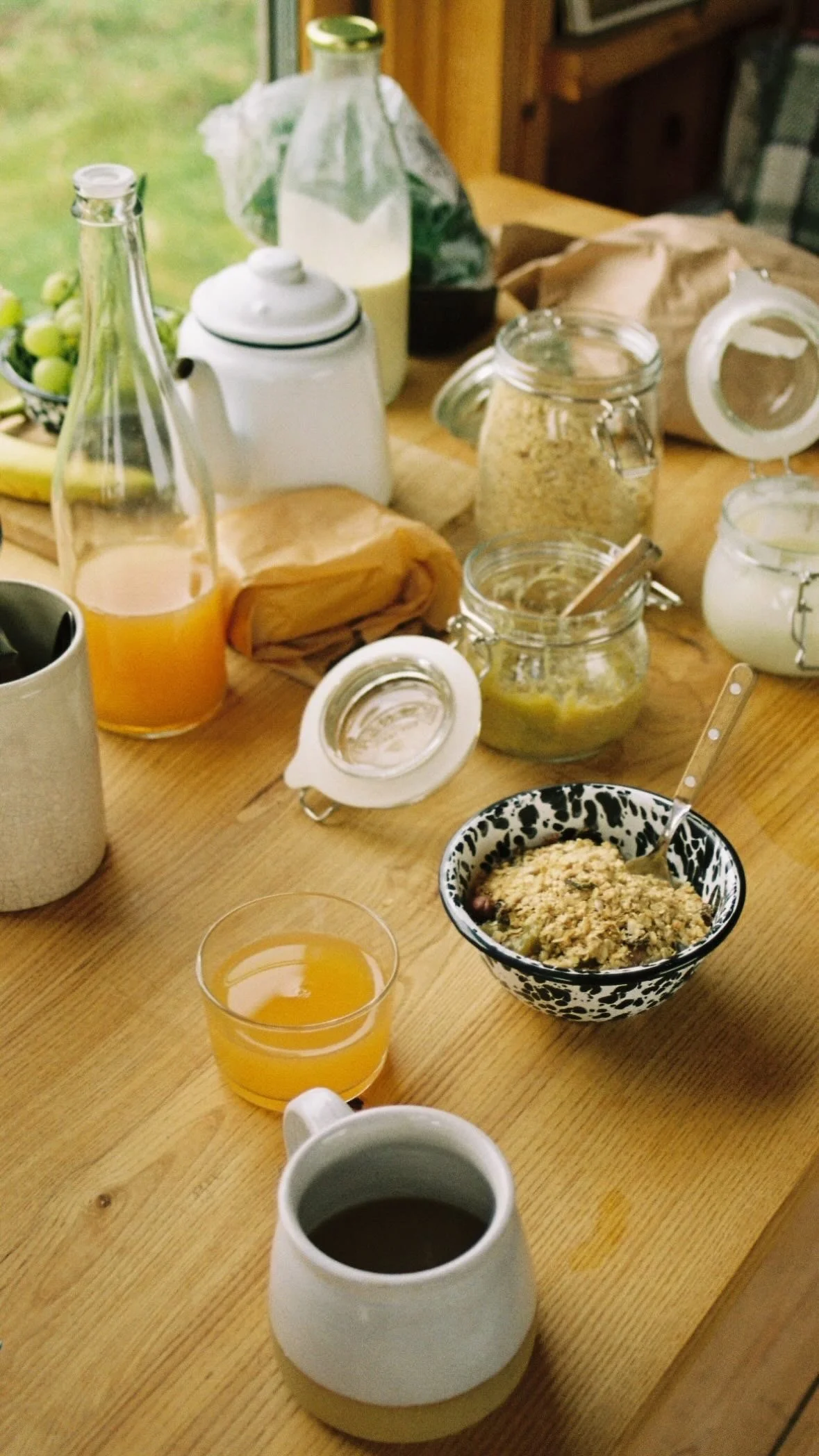 A breakfast table laid out in an offgrid hut. Hand thrown pottery mug of coffee, speckled enamel bowl of homemade granola and homemade Diggers apple juice