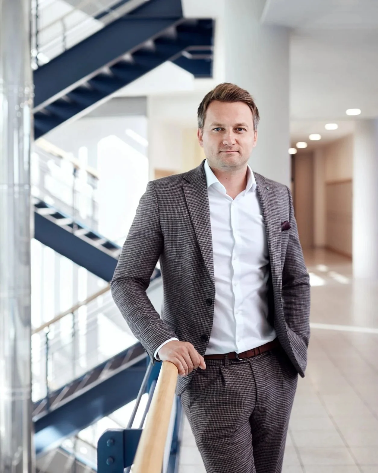 Ein Mann in Business-Kleidung steht in einem modernen Gebäude mit einer Treppe im Hintergrund.
