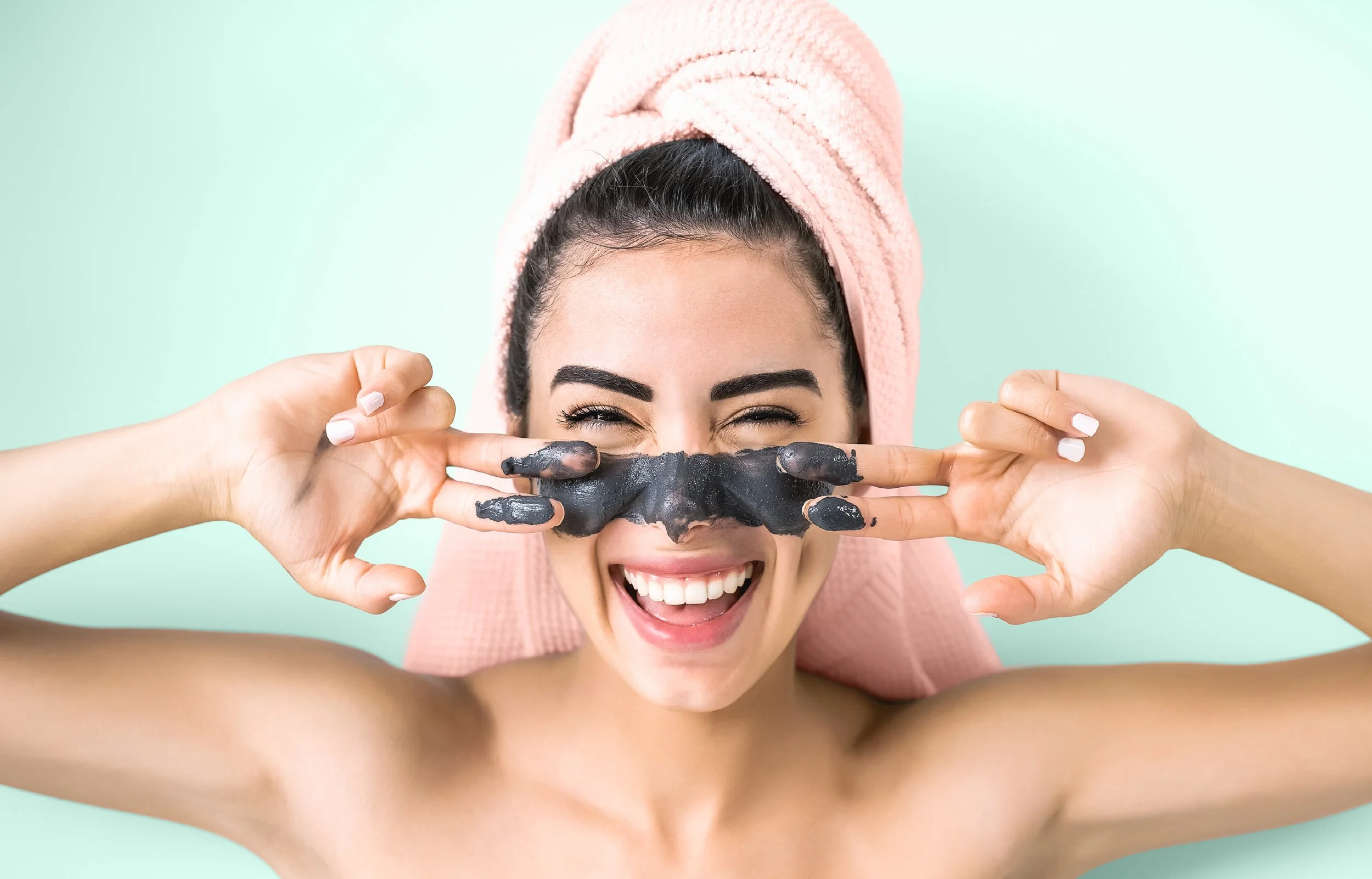 Woman with a pink towel wrapped around her hair smiling and holding her hands like glasses near her eyes, with a black mud mask on her nose, against a light mint green background.