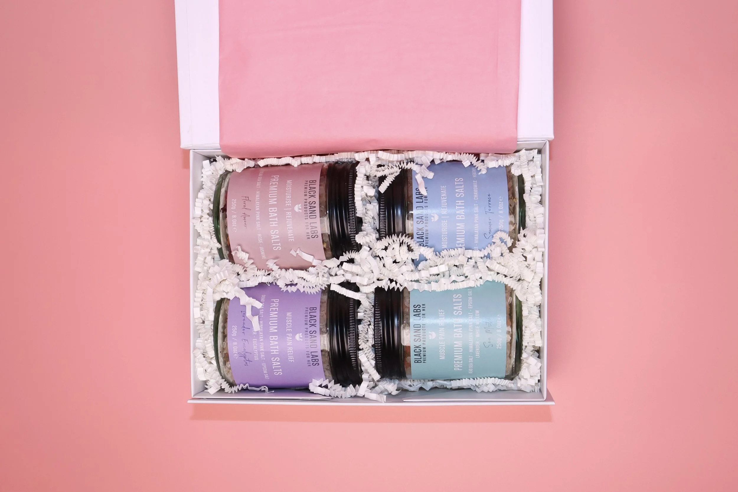Open gift box with four jars of bath salts and scrub on pink background.