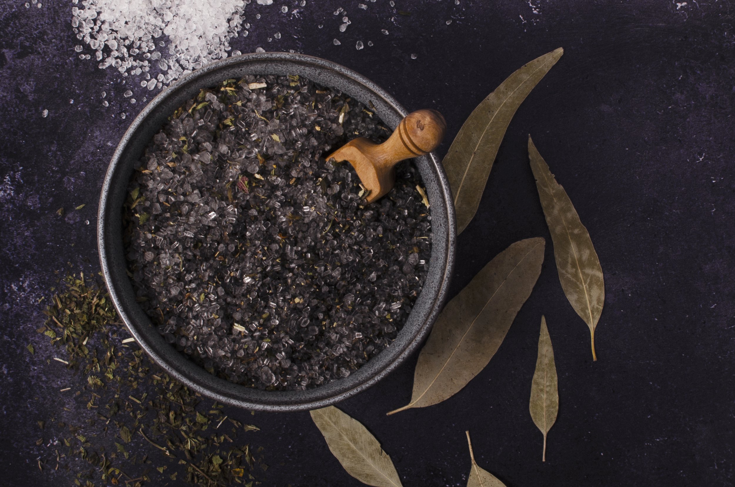 A black bowl filled with dried lavender buds, a wooden pestle resting on the edge, with dried bay leaves nearby, and scattered dried herbs and salt around the bowl on a dark surface.