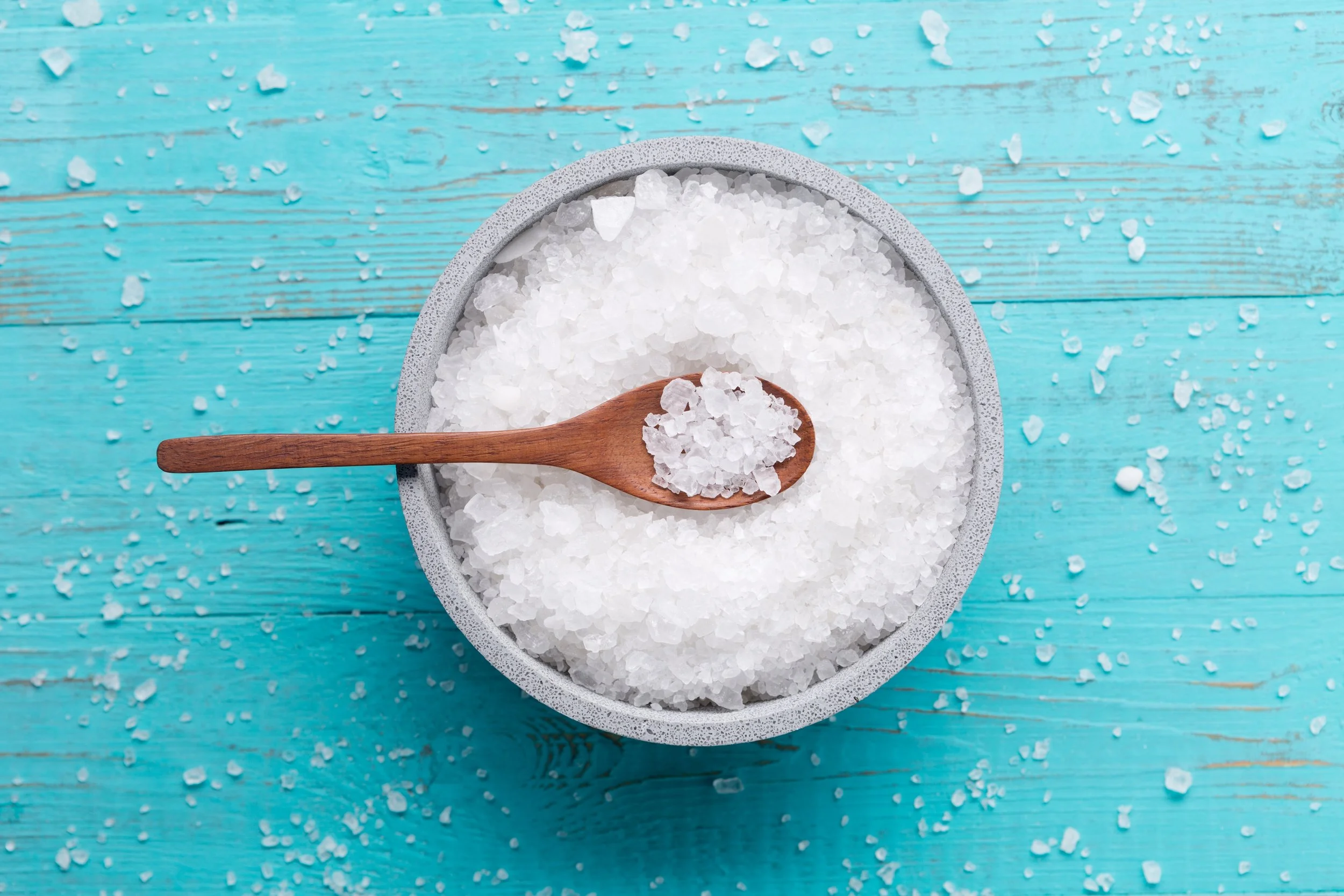 A wooden spoon resting on a bowl filled with coarse white sea salt, on a turquoise wooden surface with scattered salt crystals.