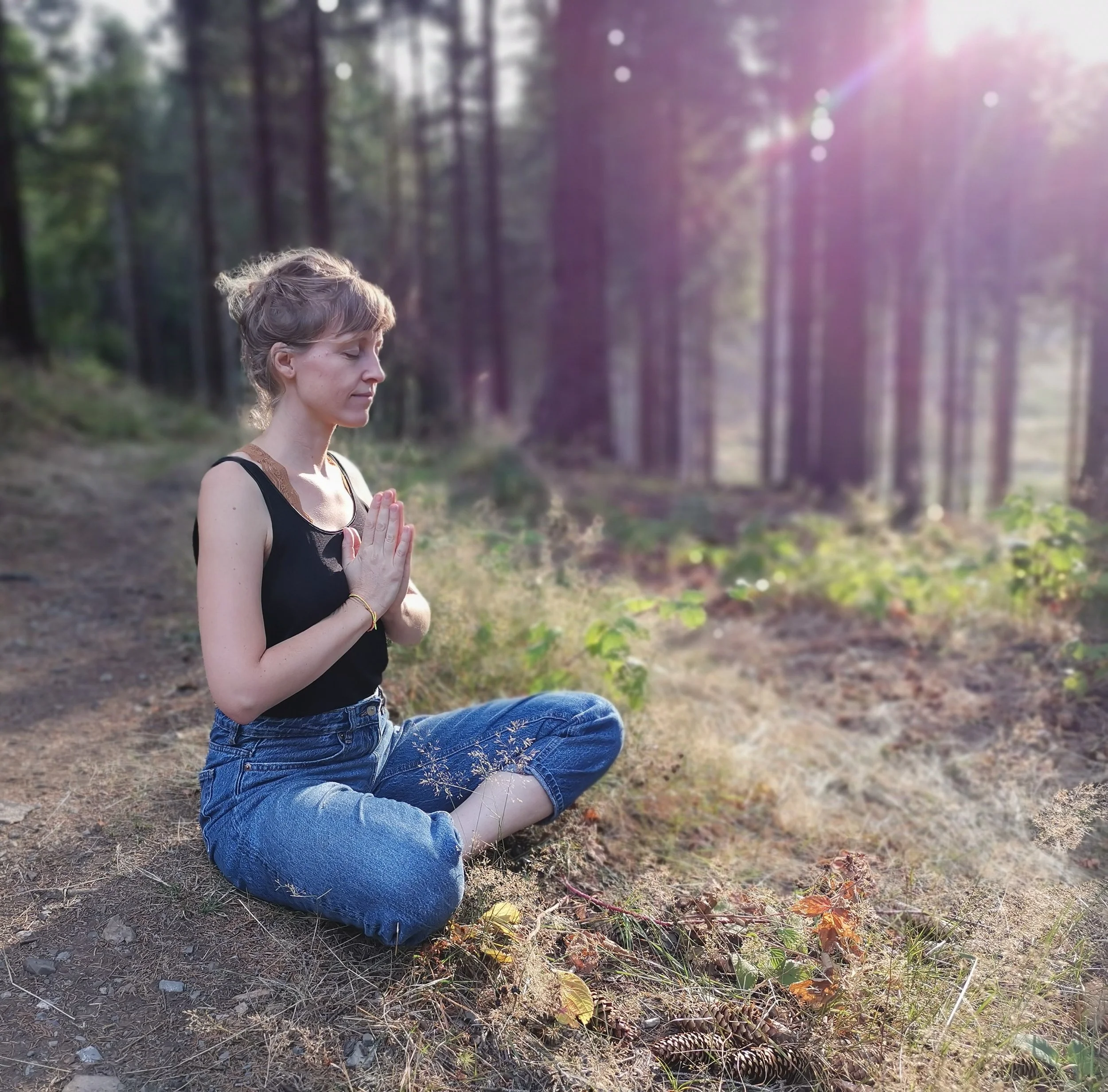 Eine Frau im Wald sitzt in Meditationshaltung mit Sonnenlicht im Hintergrund
