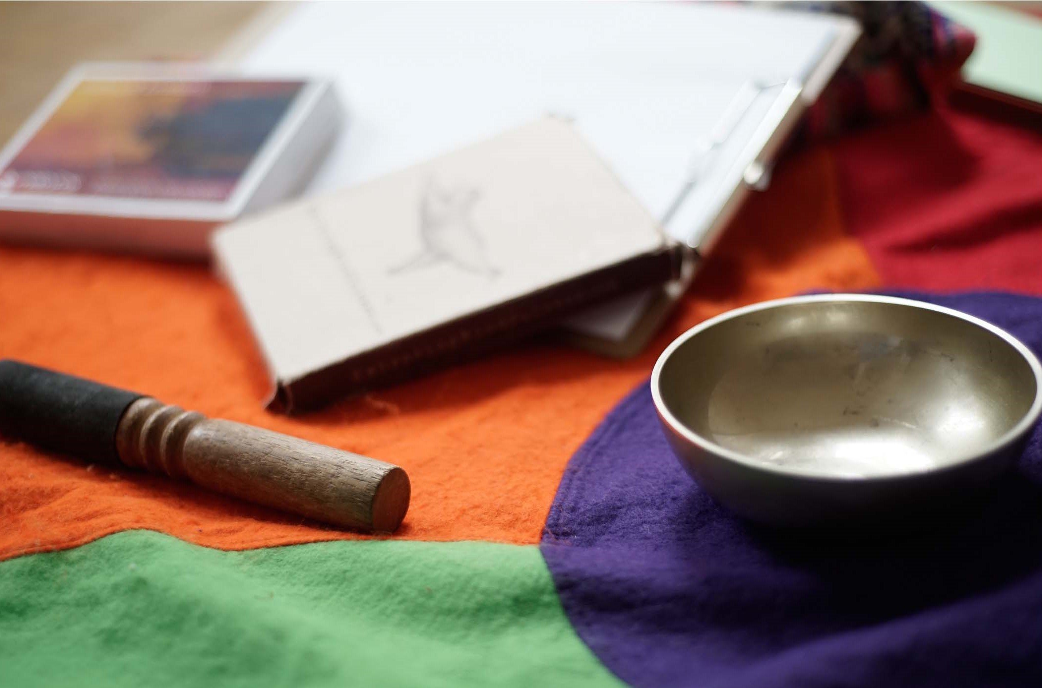 Eine Klangschale, ein Klemmbrett, Bildkarten, alle auf einem bunten Teppich in Farben Orange, Grün, Lila, Rot.