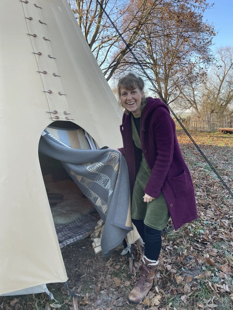 Lachende Frau in winterlicher Kleidung öffnet den Eingang zu einem Tipi. Im Hintergrund sind Bäume und blauer Himmel zu sehen.