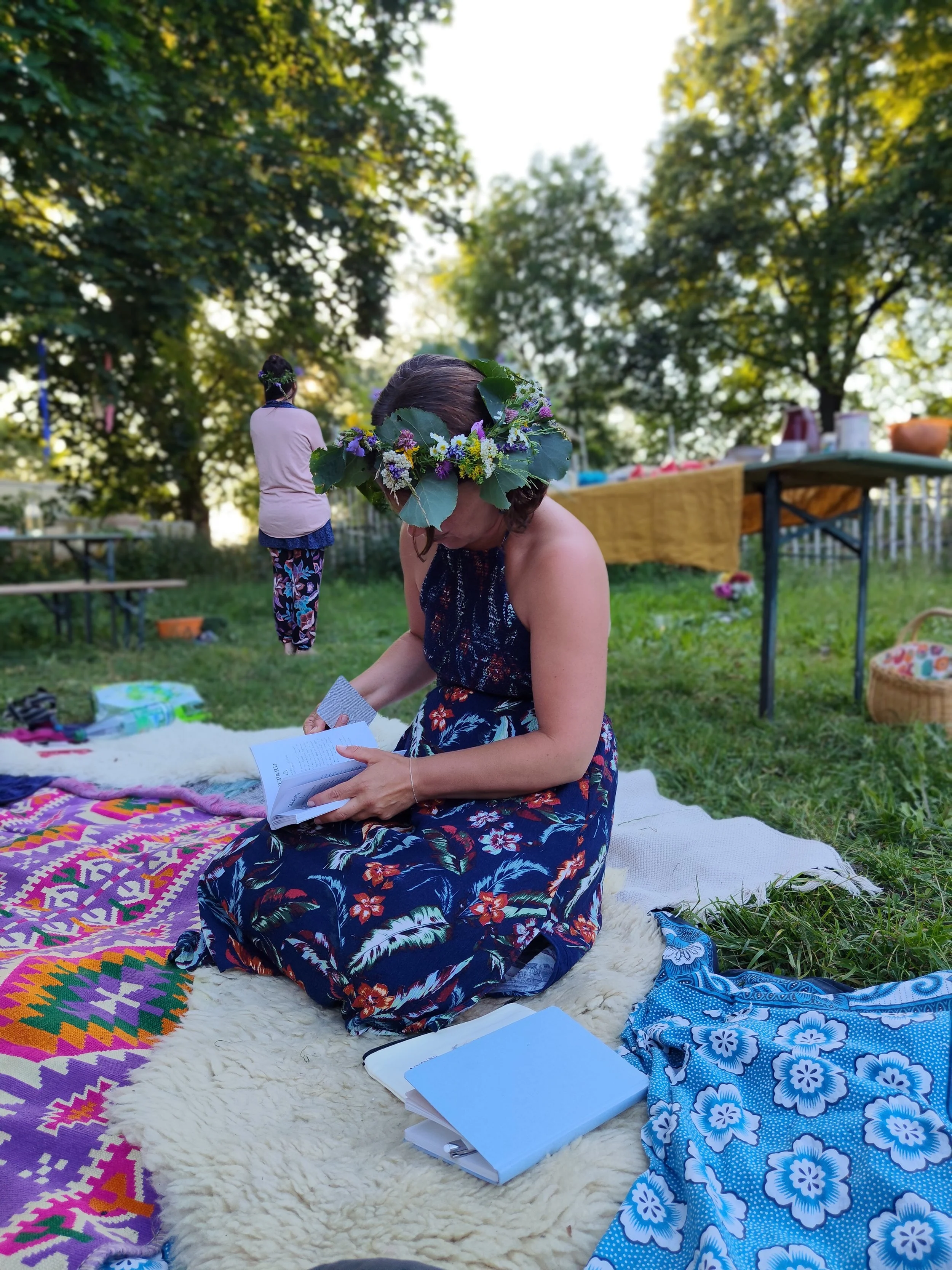 Sommerlich gekleidete Frau mit Blumenschmuck im Haar sitzt auf schön gestalteter Unterlage in einem Garten. Sie wirkt vertieft in ein Büchlein. Im Hintergrund steht eine andere Frau mit Blumenschmuck.