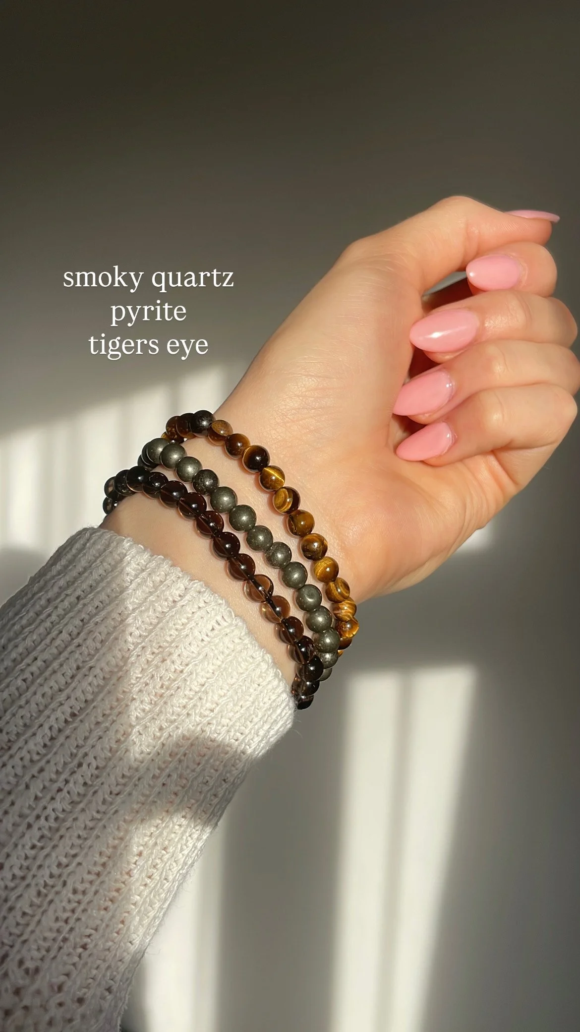 Close-up of a hand wearing three beaded bracelets made of smoky quartz, pyrite, and tiger's eye stones, with sunlight and shadows creating a warm glow.