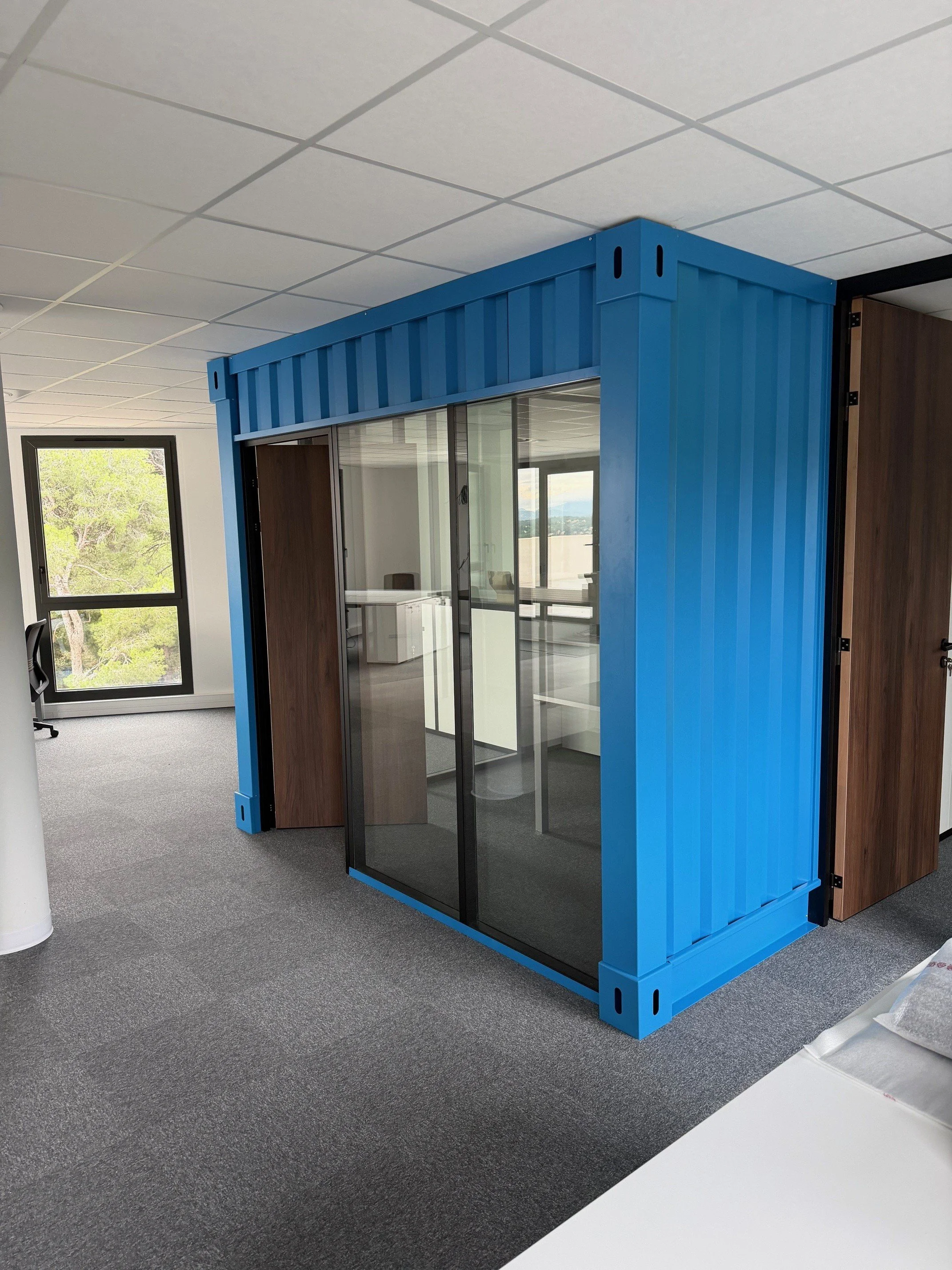 Un bureau atypique construit à l'intérieur d'un conteneur bleu avec des cloisons vitrées.